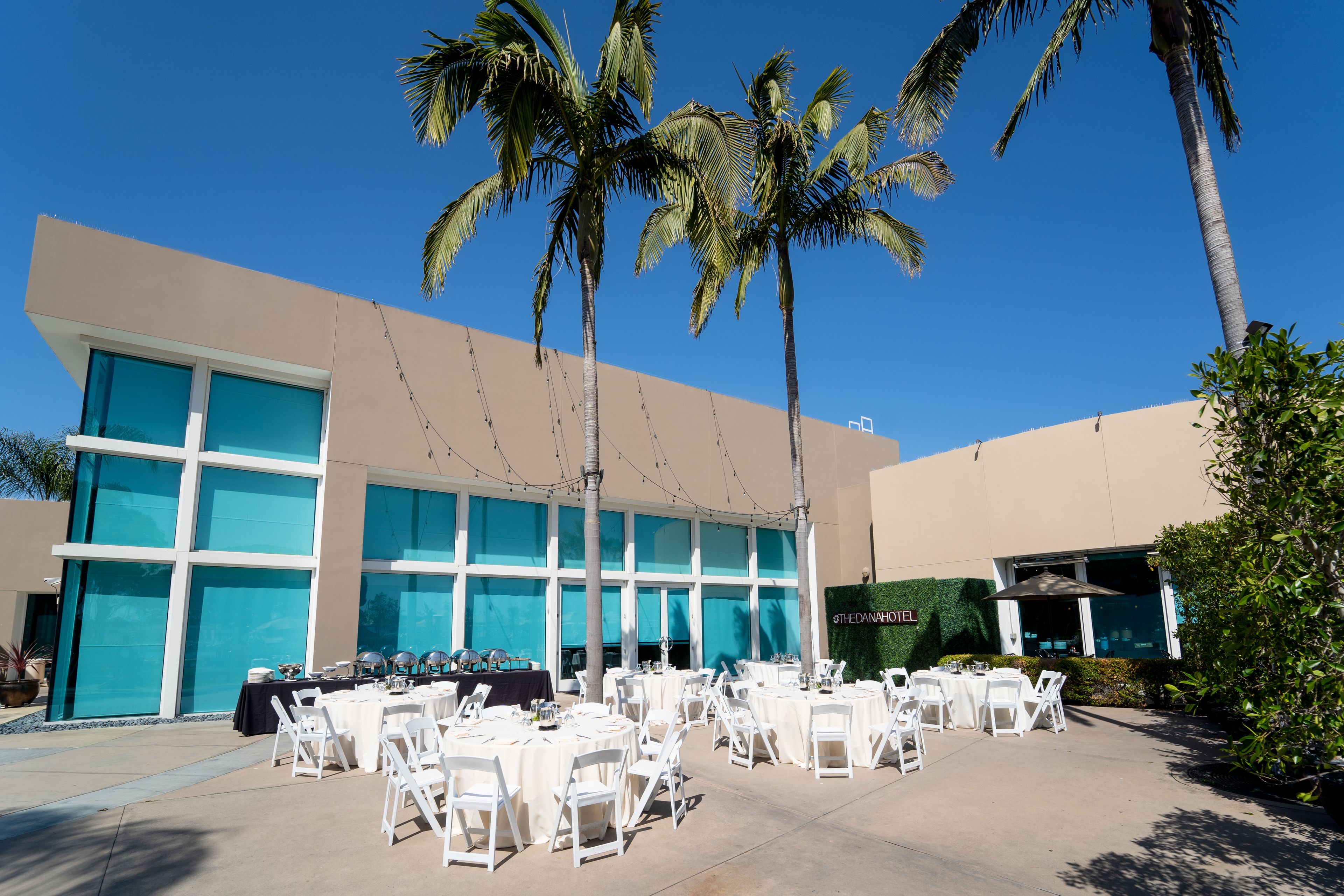 An outdoor dining area with white tables and chairs, palm trees, and a building with large windows and a sign that reads "THE DANAHOTEL".