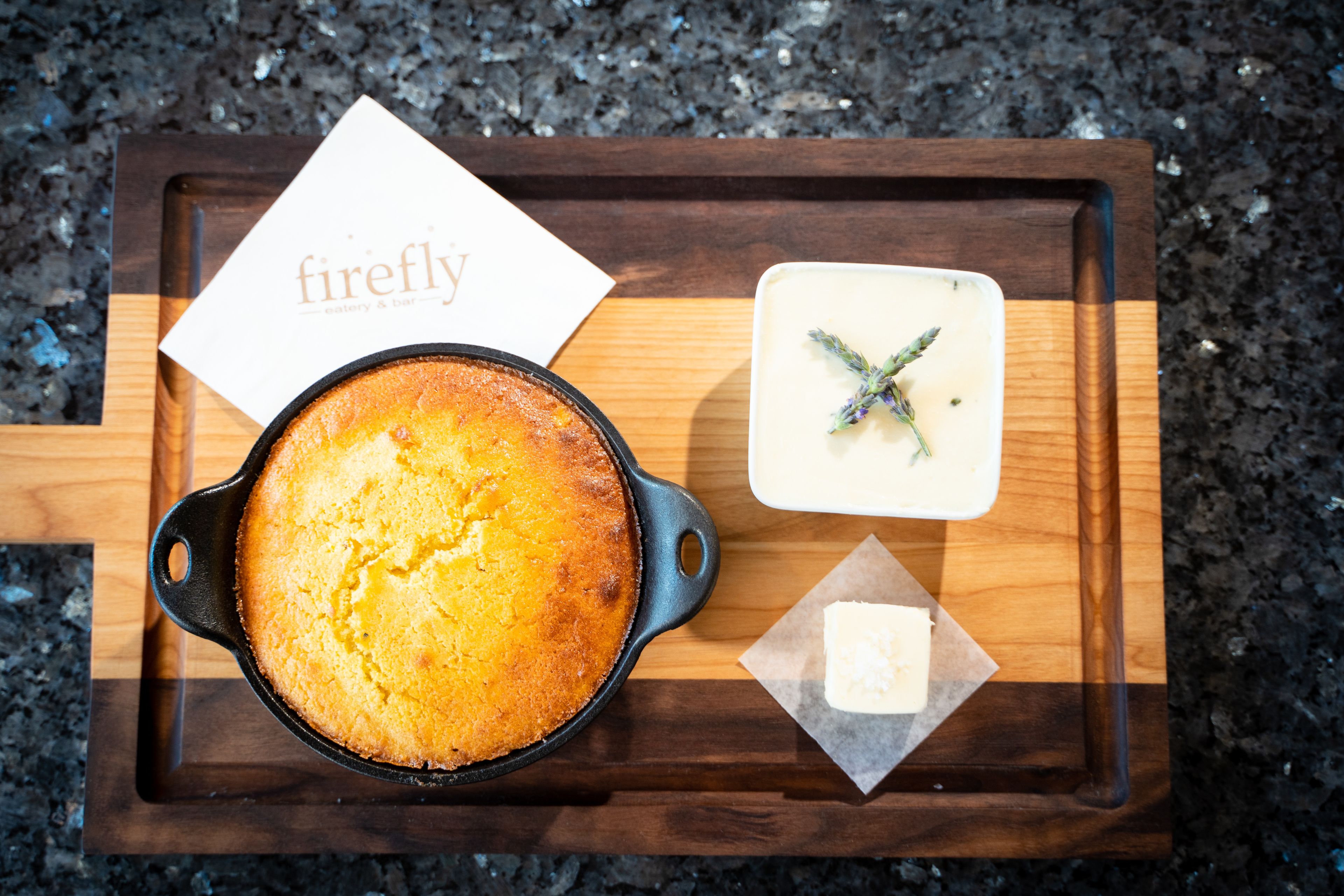 A cast iron skillet with a baked dish, a small dish of white sauce with herbs, and a piece of butter on a wooden tray.