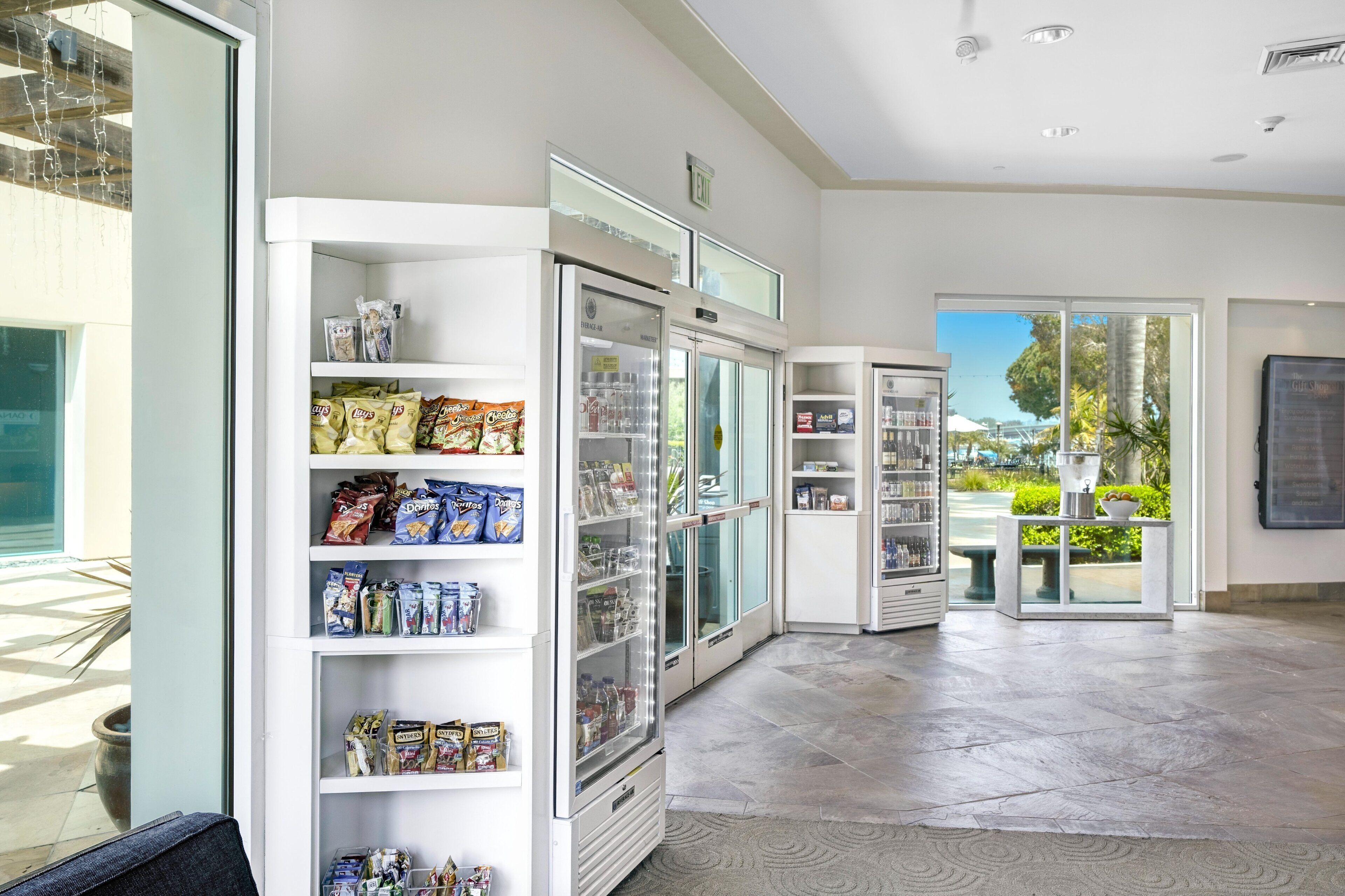 Un moderno vestíbulo de hotel con un expositor refrigerado, una zona de estar con vistas al exterior y un mostrador de recepción.