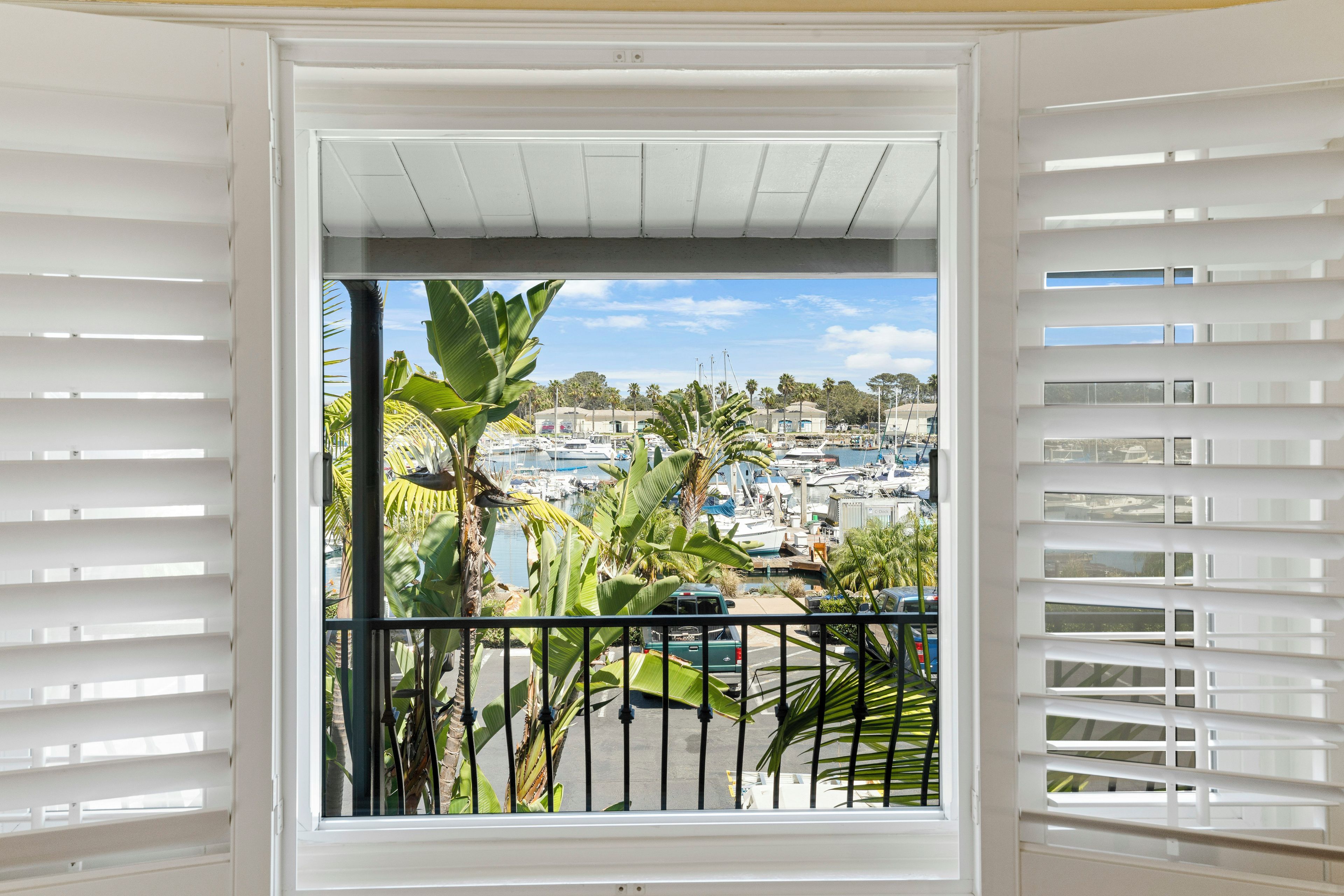 Un balcón con contraventanas blancas, vista a un puerto deportivo con barcos y plantas tropicales.