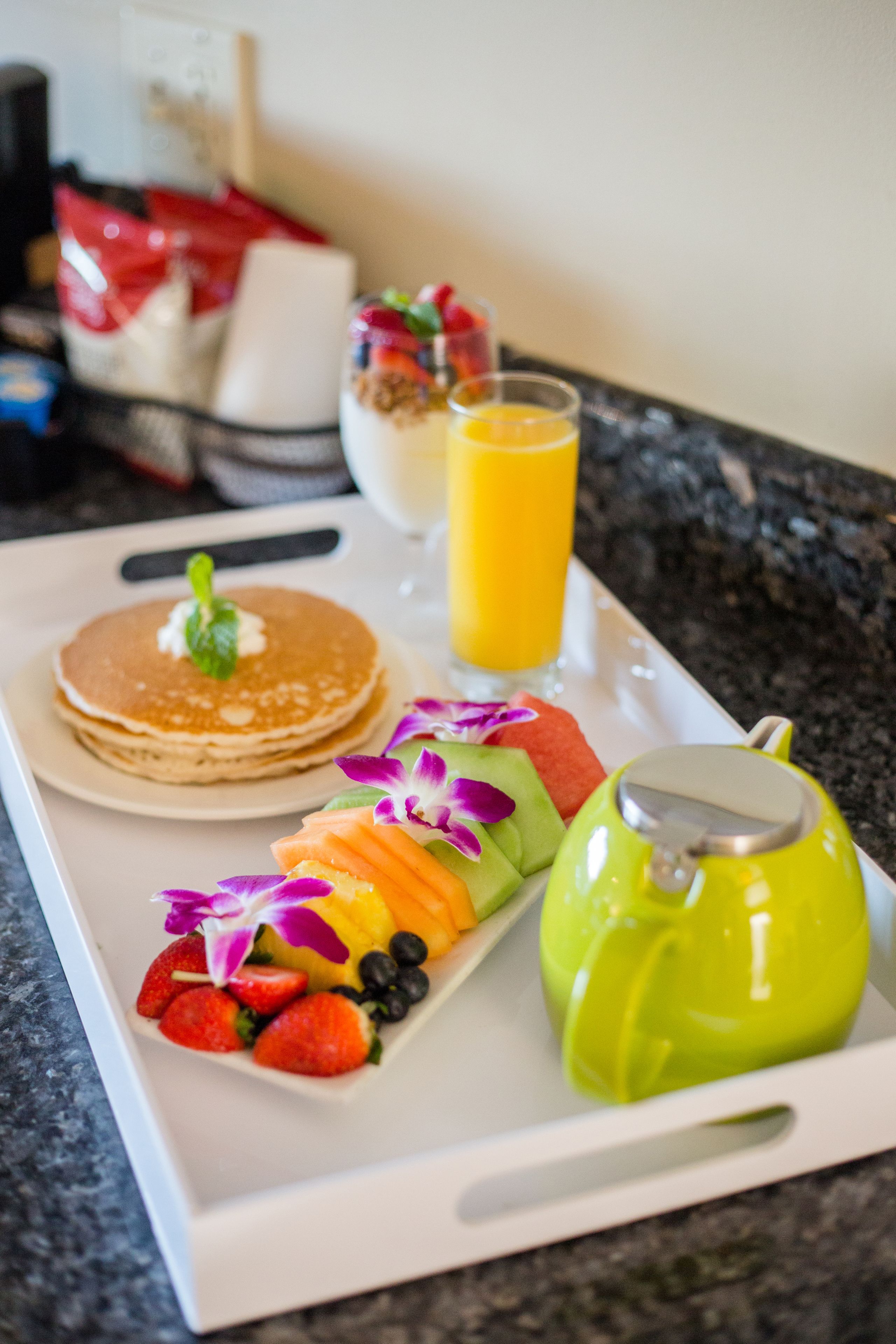 A breakfast tray with pancakes, fruit, a glass of orange juice, and a green teapot.