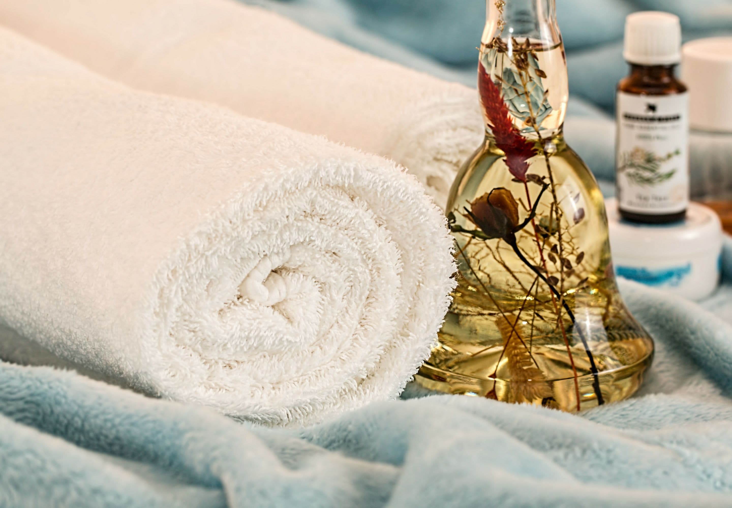 A rolled white towel, a glass bottle with a flower, and a small bottle with a label on a textured blue surface.