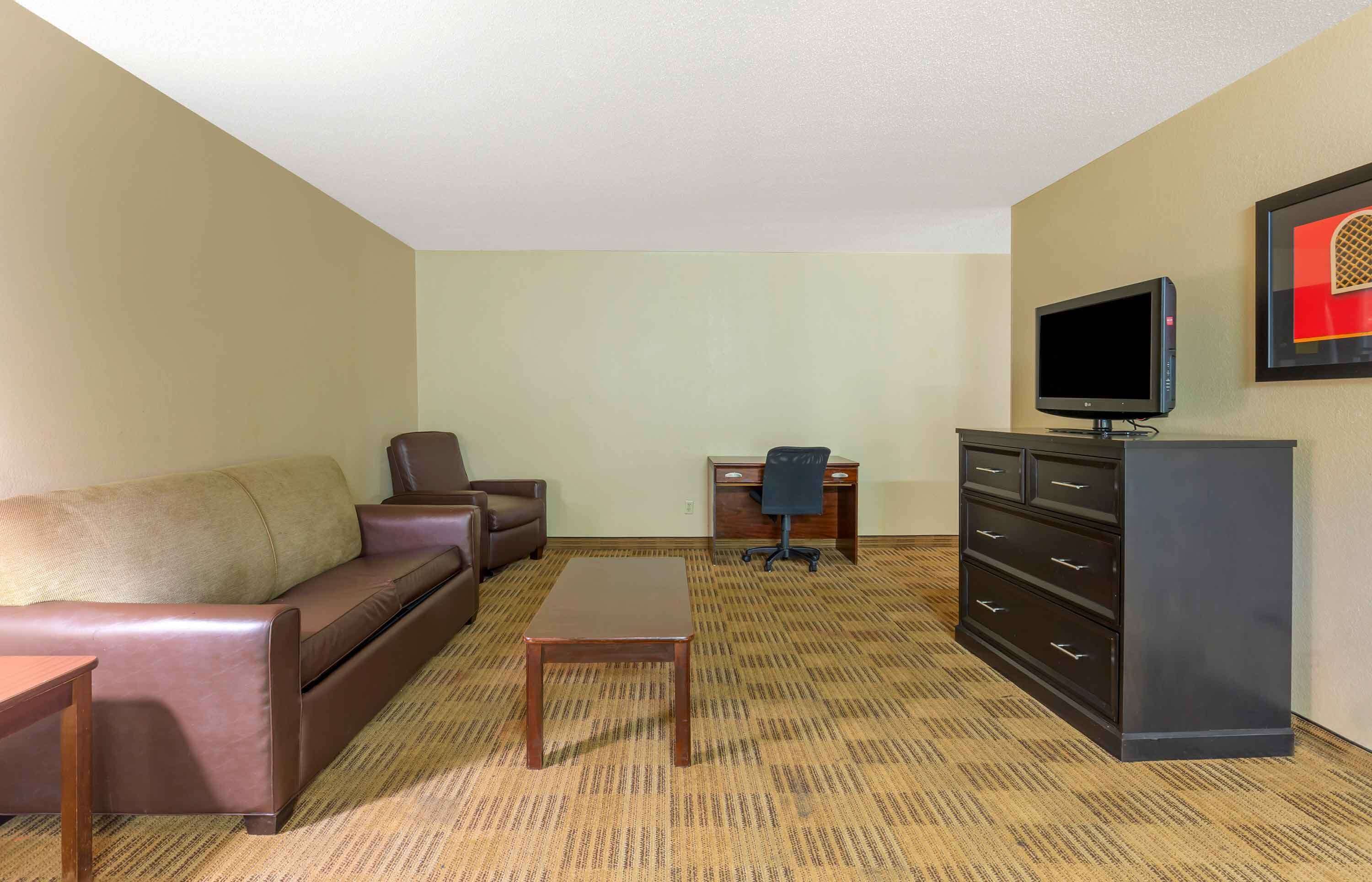A hotel room with a brown leather sofa, a wooden coffee table, a black office chair, a desk with a computer, a black dresser, and a flat-screen TV mounted on the wall.