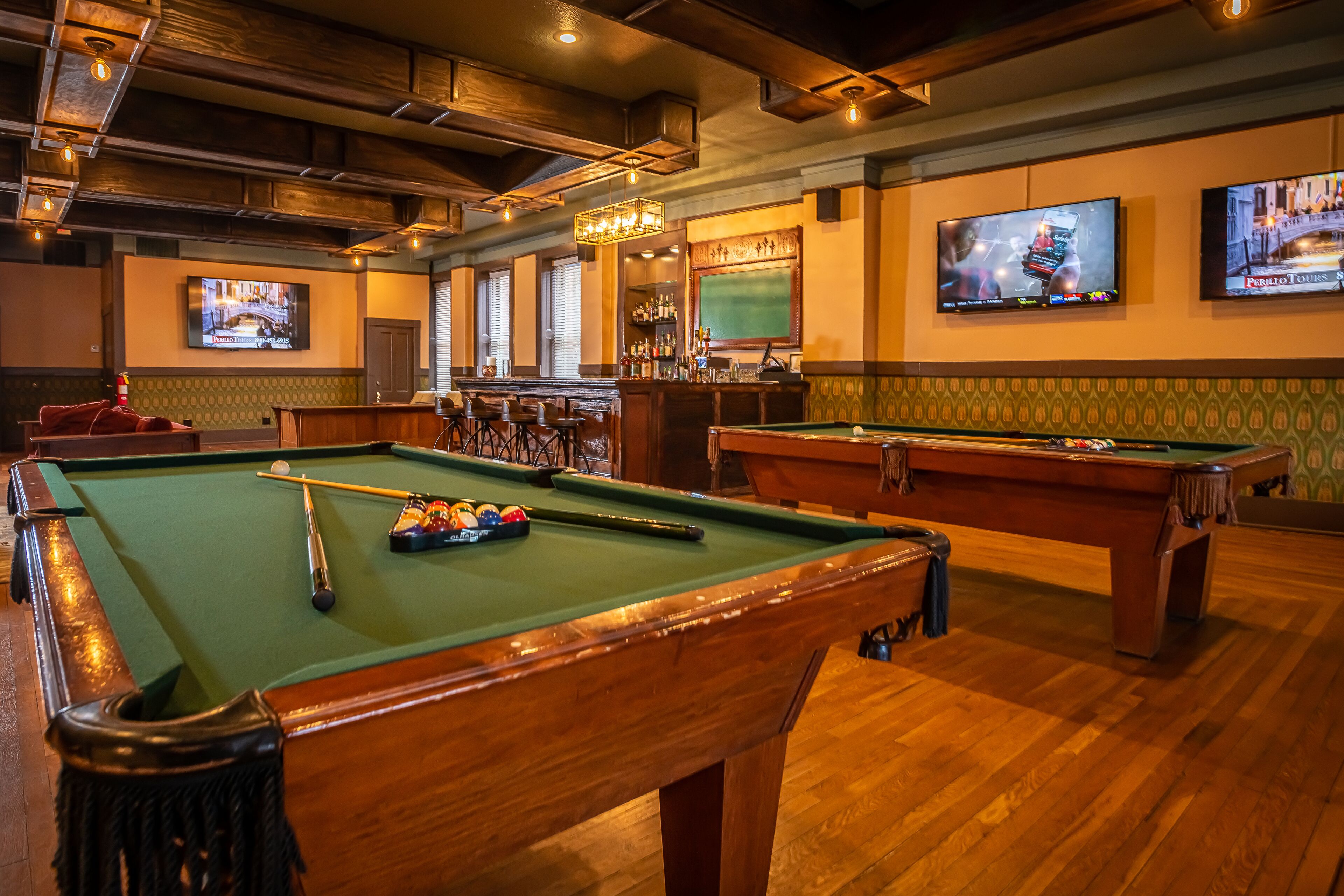 A pool table with billiard balls, a bar area with a television, and a green chalkboard on the wall.