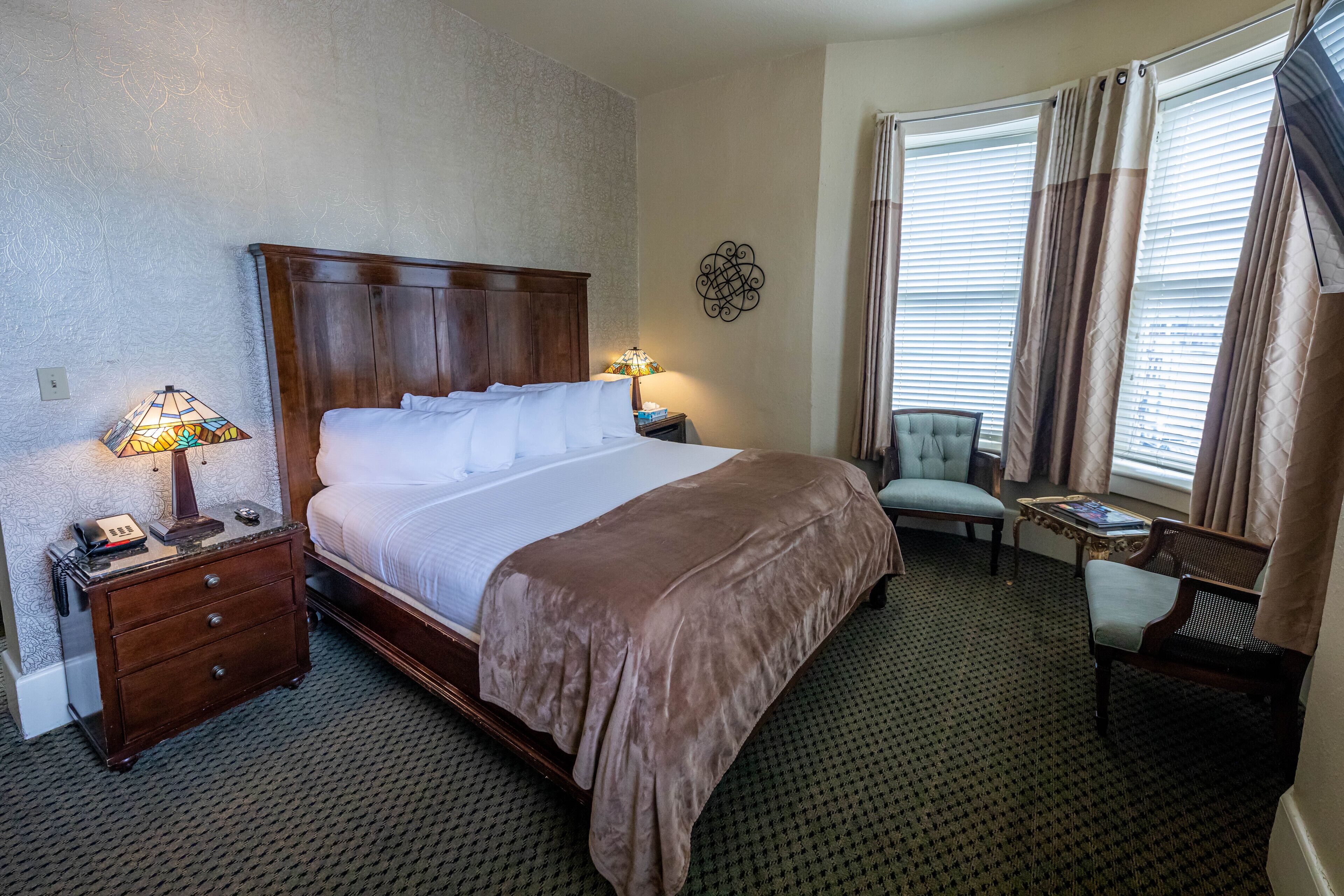 Een hotelkamer met een groot bed, een nachtkastje met een lamp, een stoel, een klein tafeltje en een raam met gordijnen.
