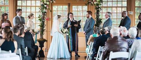 A wedding ceremony in a room with large windows, wooden floors, and a floral arch.