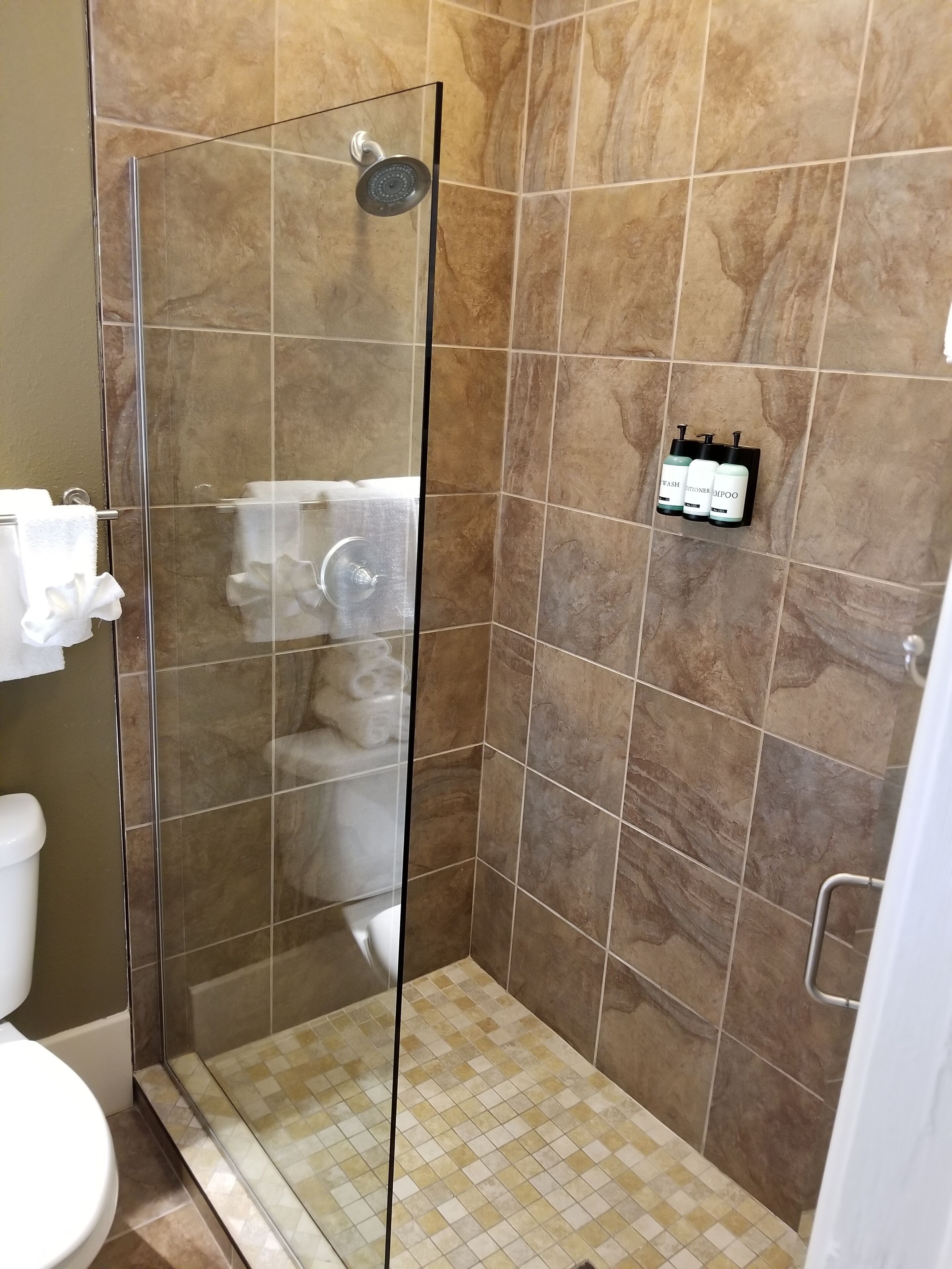 A shower area with a glass door, beige tiles, and a small shelf with toiletries.