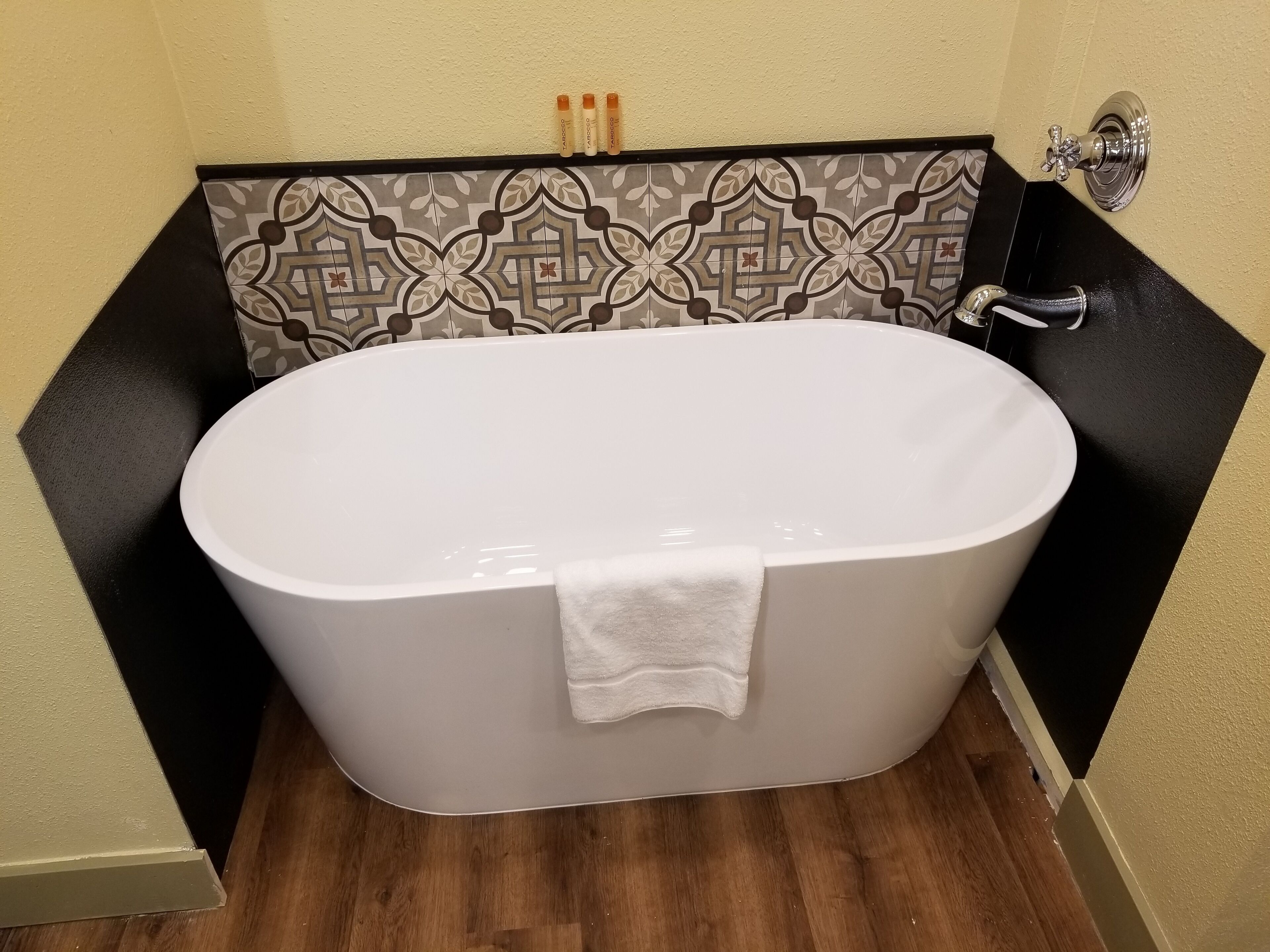 A white freestanding bathtub with a white towel, a patterned tile wall, and a black countertop.