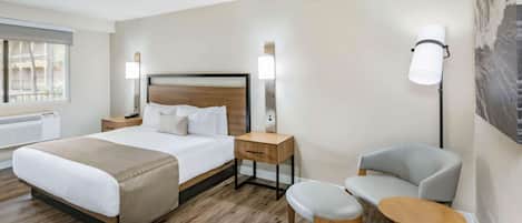 A hotel room with a large bed, bedside tables, a chair, a small table, and a wall-mounted lamp.