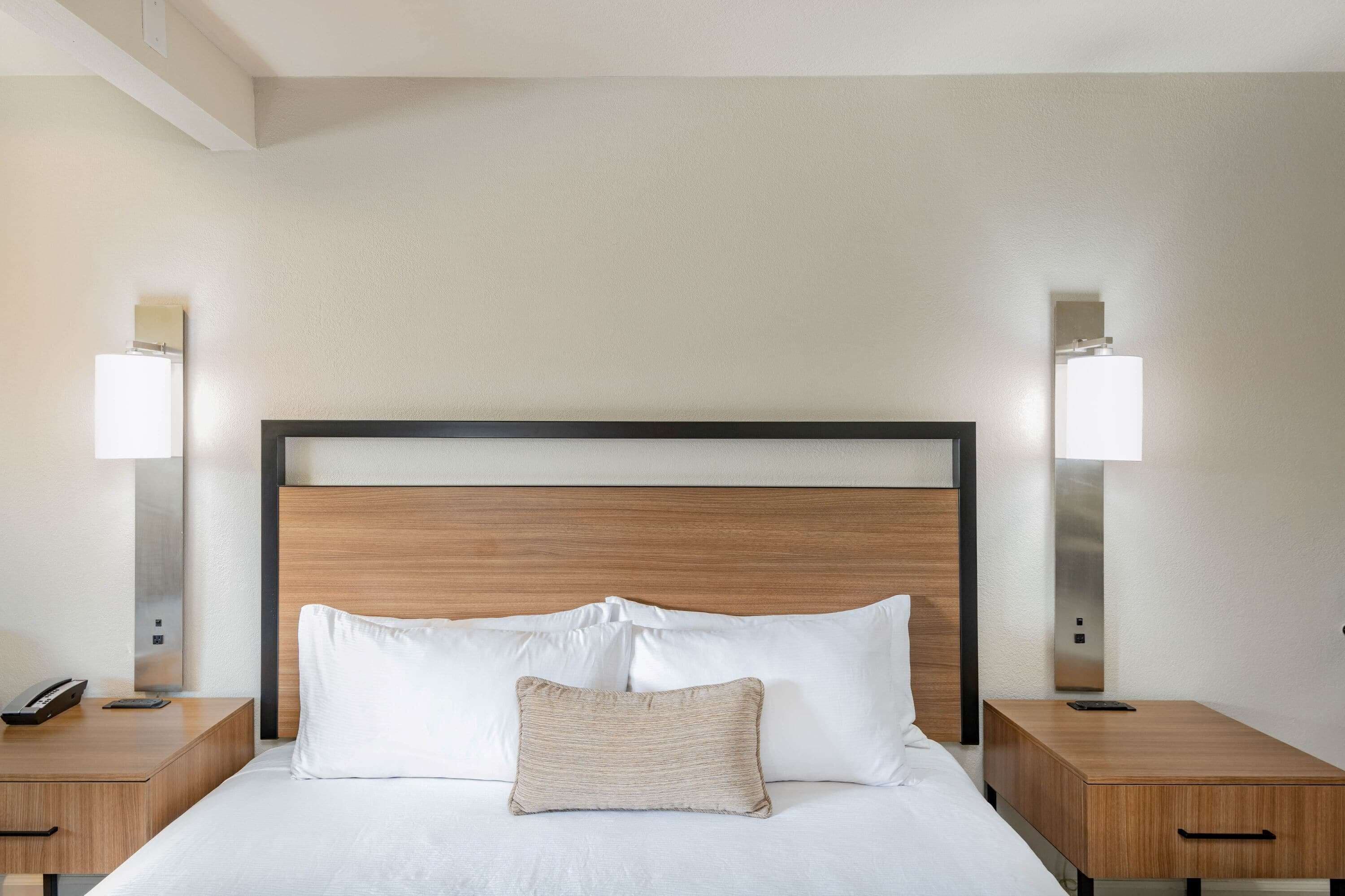 Habitación de hotel con cabecera de madera, ropa de cama blanca, almohada decorativa color beige y lámparas de noche.