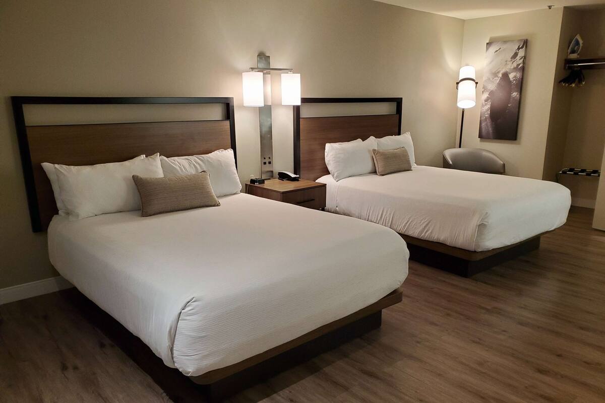 Habitación de hotel con dos camas, una mesita de noche con lámpara, una silla y una repisa en la pared.