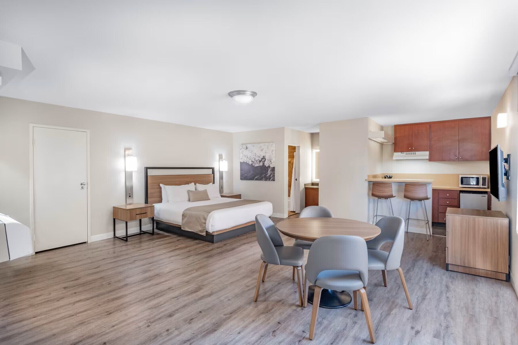 A hotel room with a bed, bedside tables, a desk, a dining table with chairs, a kitchen area, and a TV.