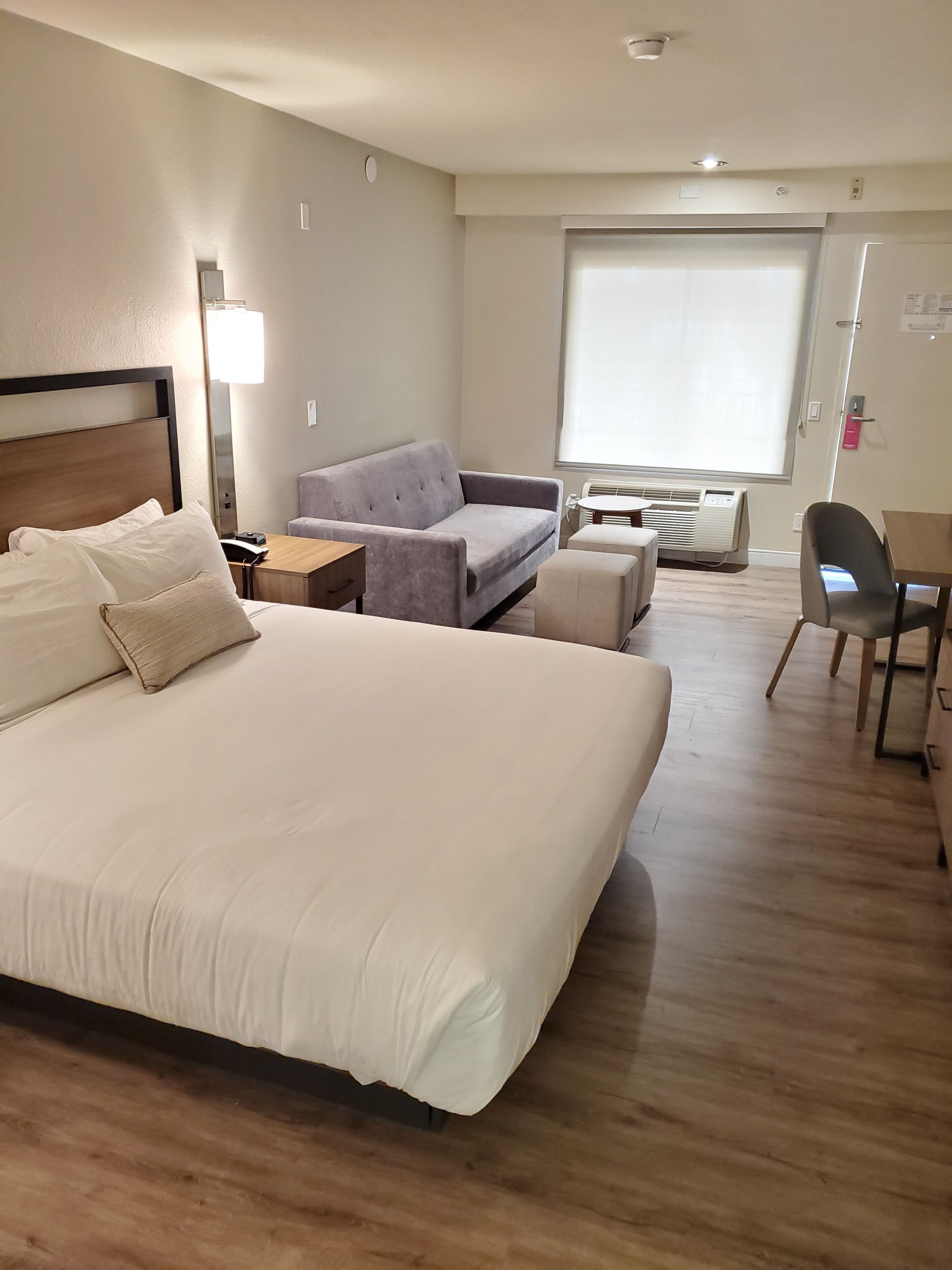 A hotel room with a large bed, a grey sofa, a small table, and a chair.