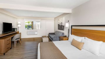 A hotel room with a bed, a desk, a television, a sofa, and a window with a view of greenery.