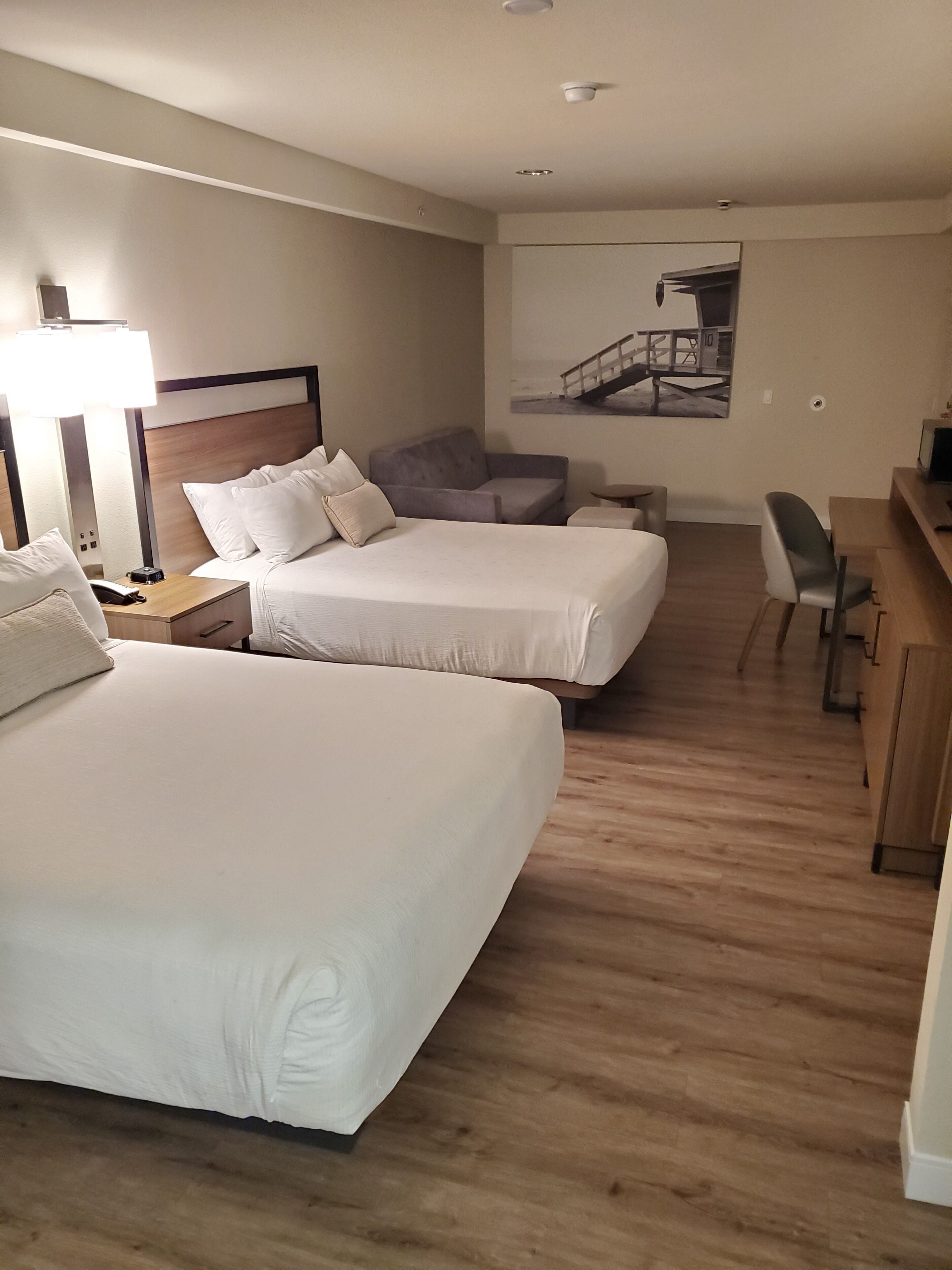 A hotel room with two beds, a desk, a chair, a sofa, and a lamp.