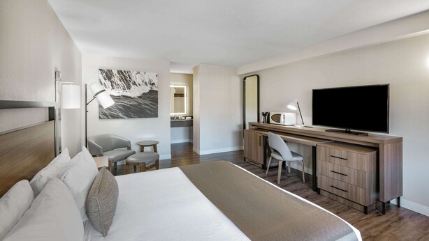 A modern hotel room with a large bed, a desk with a flat-screen TV, a chair, a small table, and a painting on the wall.