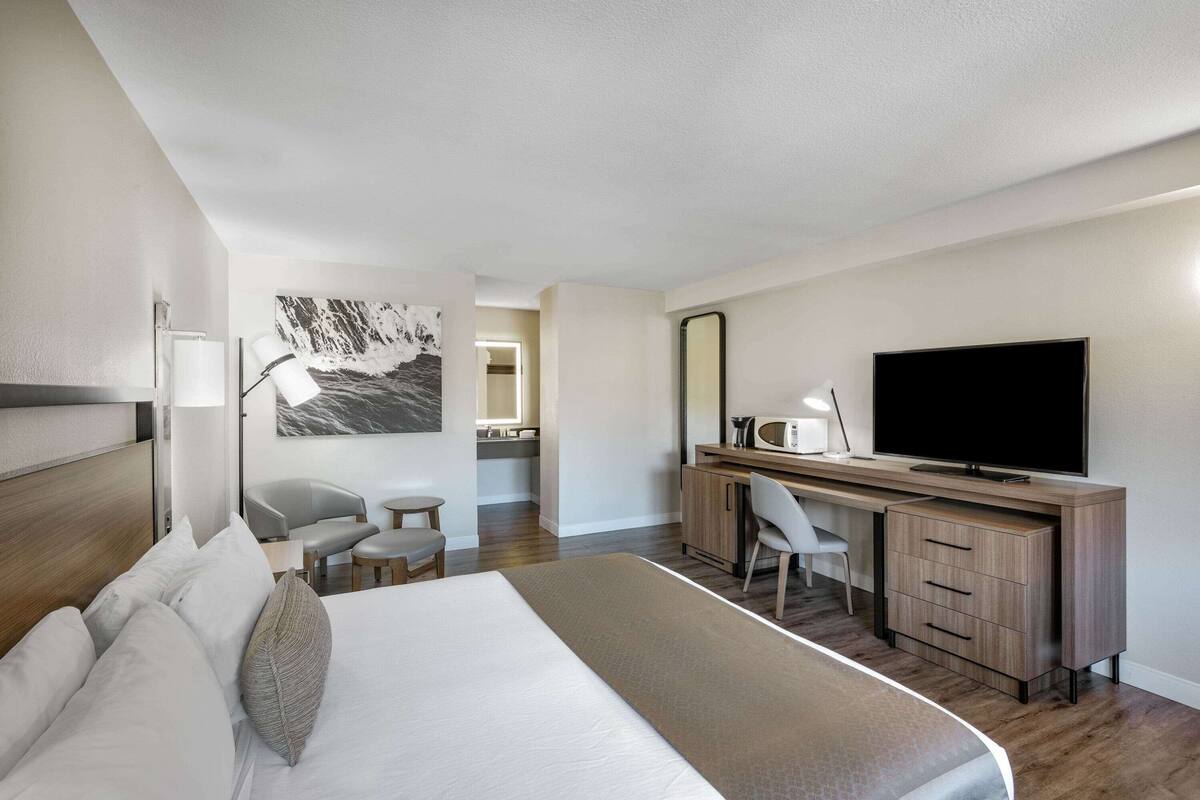 Una habitación de hotel moderna con una cama grande, un escritorio con televisión de pantalla plana, una silla, una mesita y un cuadro en la pared.