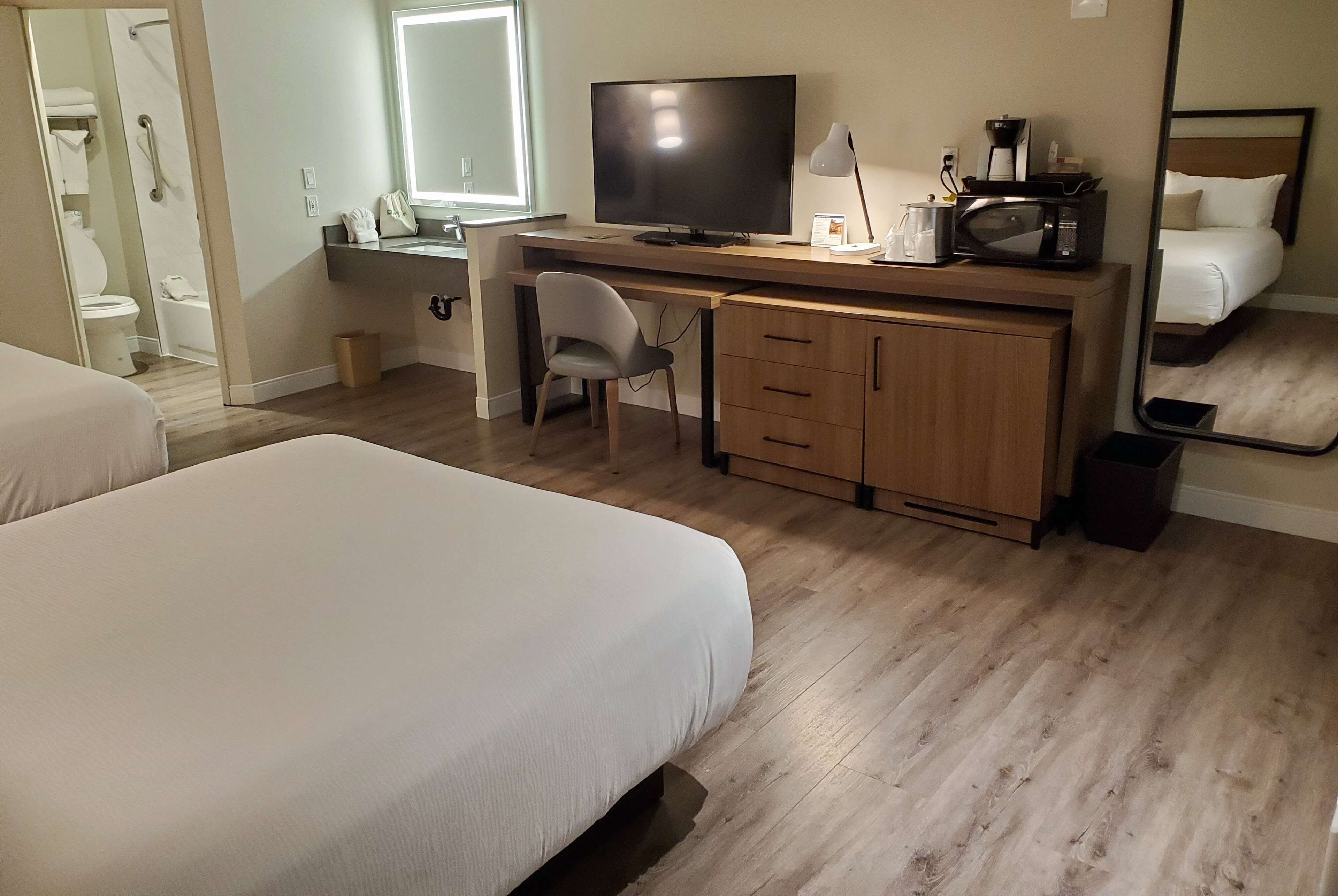 A hotel room with a bed, a desk with a TV, a chair, and a large mirror.