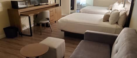 A modern hotel room with a wooden desk, a television, a bed with pillows, a sofa, and a small round table.