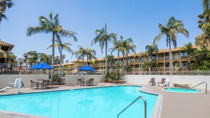 A swimming pool with lounge chairs and umbrellas, surrounded by palm trees and a hotel building.