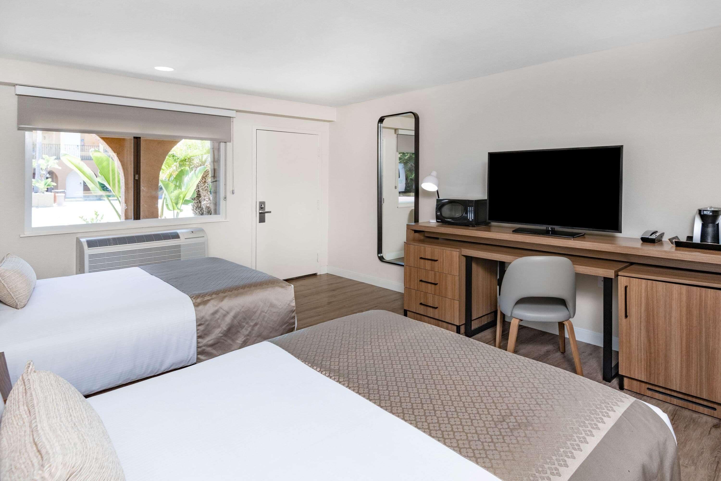 Habitación de hotel con dos camas, un escritorio con televisor, un espejo, una silla y una ventana con vistas a zonas verdes.