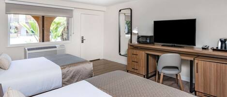 Habitación de hotel con dos camas, un escritorio con televisor, un espejo, una silla y una ventana con vistas a zonas verdes.