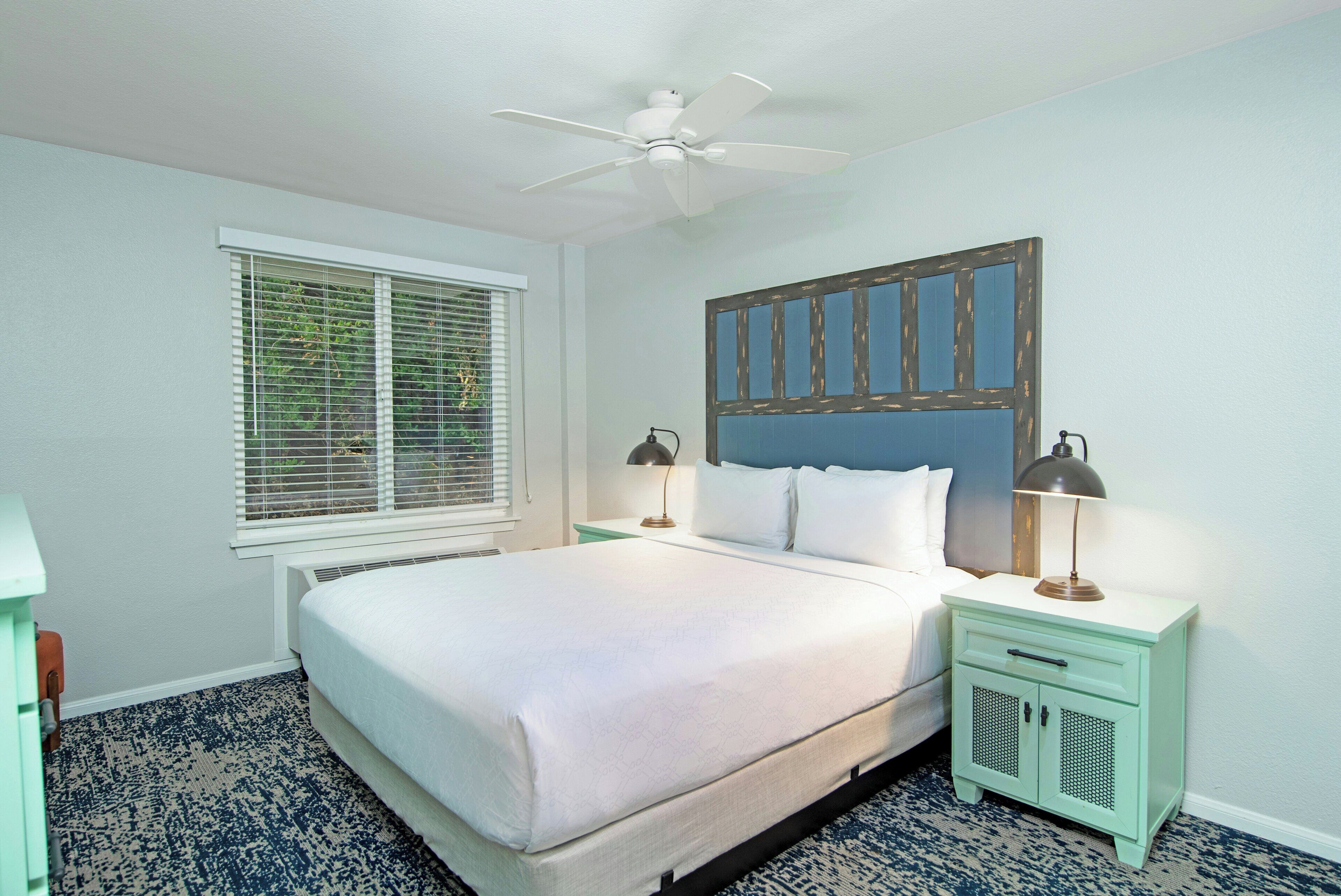 A bedroom with a bed, a ceiling fan, a window with blinds, and a nightstand with a lamp.