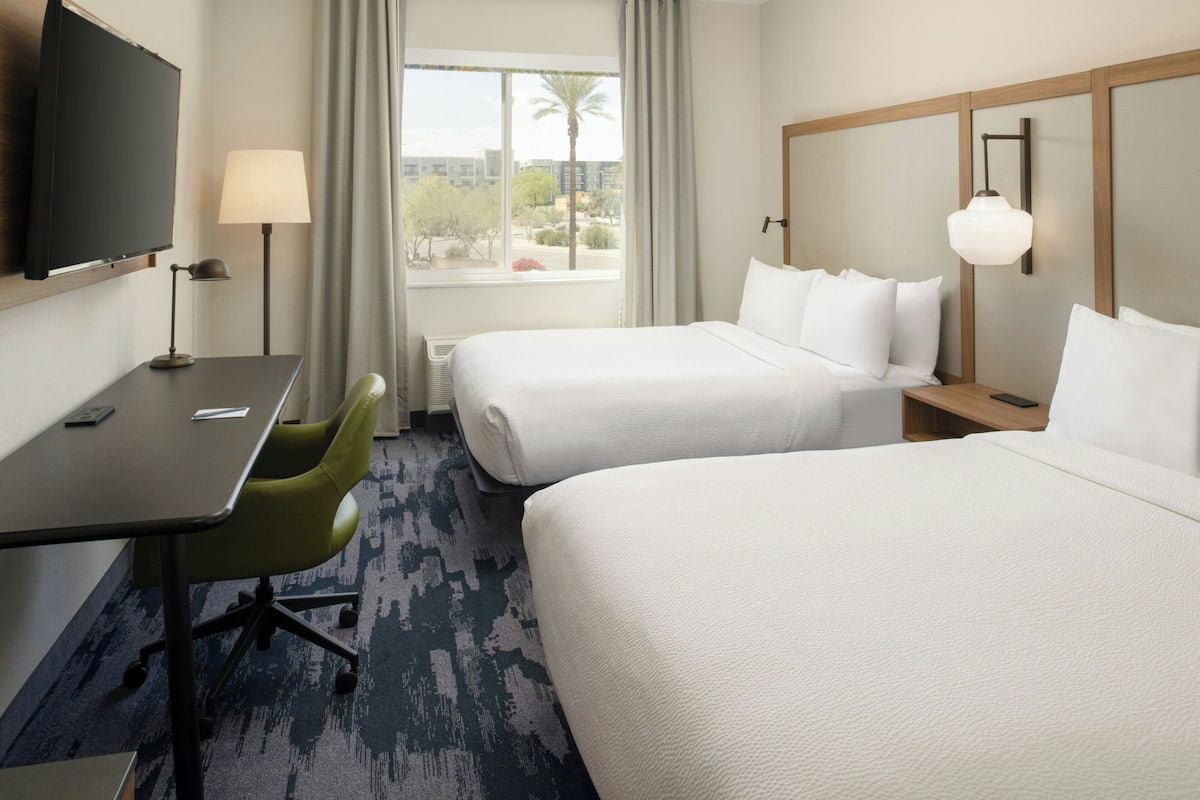 A hotel room with two beds, a desk with a chair, a TV, and a window with a view of palm trees and buildings.