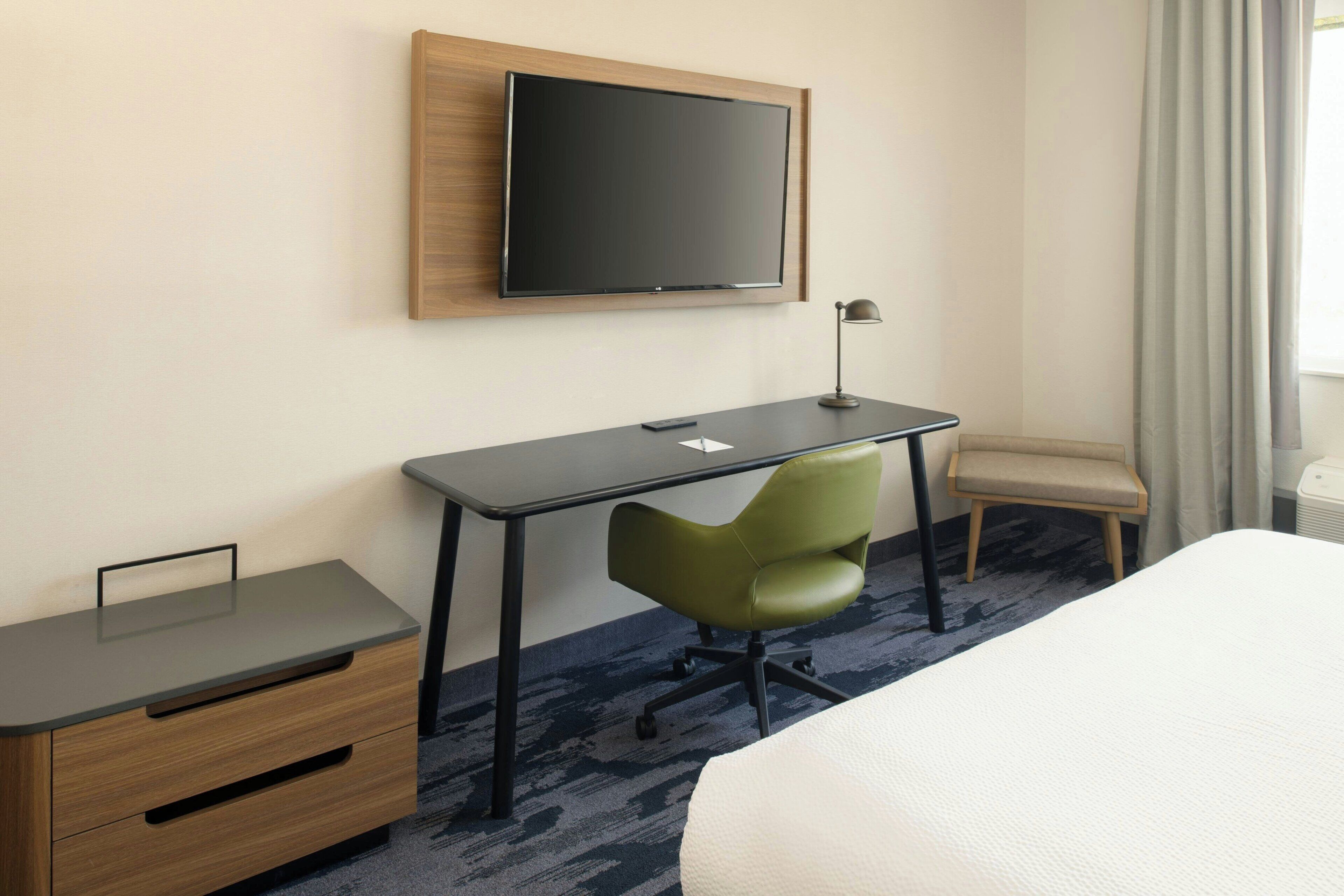 A hotel room with a bed, a desk with a chair, a television mounted on the wall, and a window with curtains.