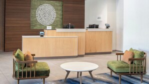 A reception area with a wooden reception desk, two green cushioned chairs, a round table, and a wall-mounted decorative piece. - Fairfield Inn & Suites by Marriott Scottsdale Old Town (Scottsdale)