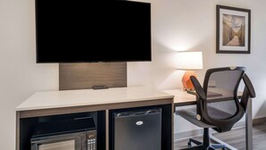 Une chambre d’hôtel équipée d’un téléviseur à écran plat fixé au mur, d’un bureau avec une lampe, d’une chaise et d’un petit réfrigérateur.