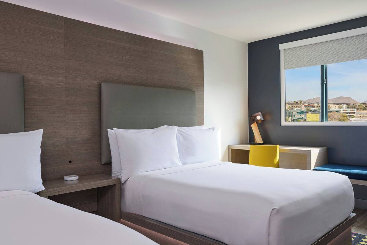 Een hotelkamer met een groot bed, een nachtkastje met een lamp, een raam met uitzicht op de stad en een zithoekje met een gele stoel.