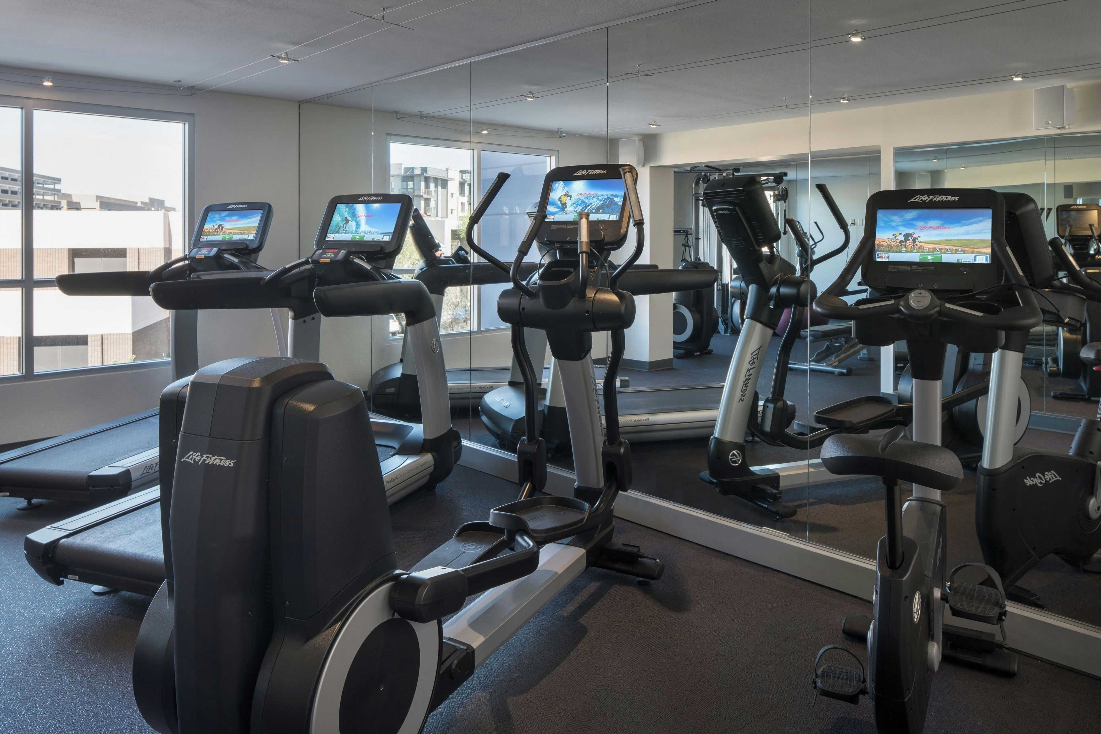Un gimnasio con múltiples cintas de correr y elípticas, espejos amplios y vistas de la ciudad a través de grandes ventanales.