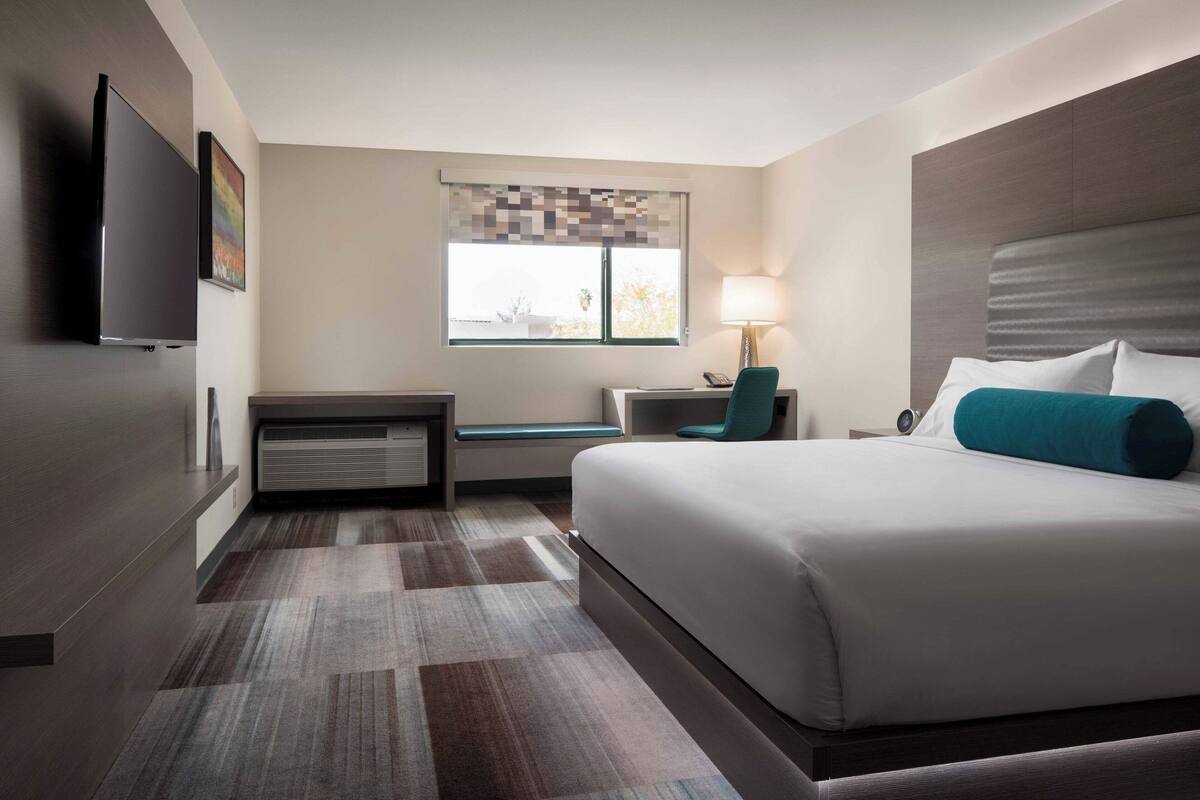 A modern hotel room with a large bed, a desk with a chair, a flat-screen TV, a window with a view, and a lamp.
