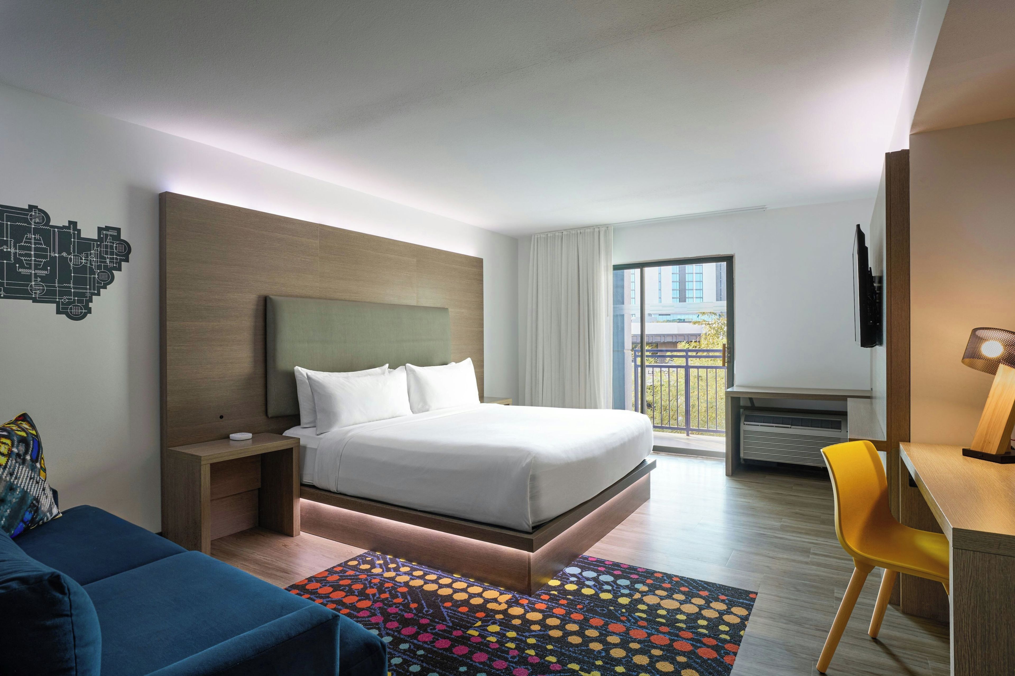 Une chambre d’hôtel avec un lit, un bureau, une chaise, un tapis coloré et une vue sur l’extérieur.