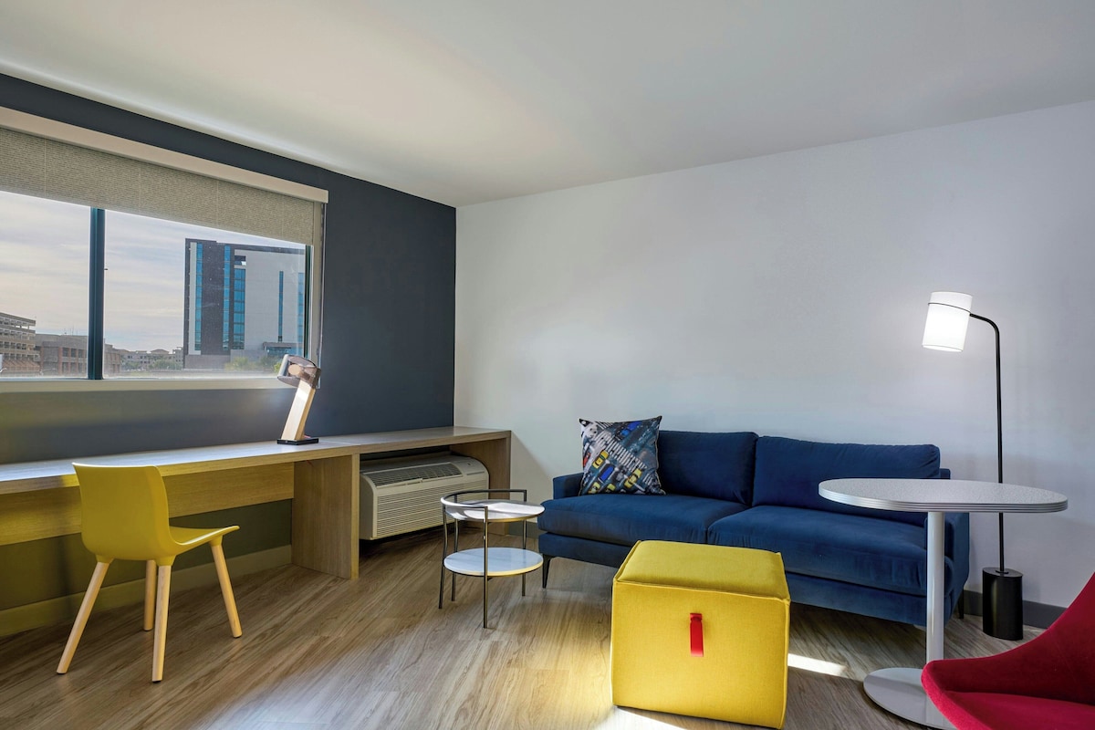 A modern living room with a desk, a blue sofa, a yellow ottoman, a red chair, and a round table.