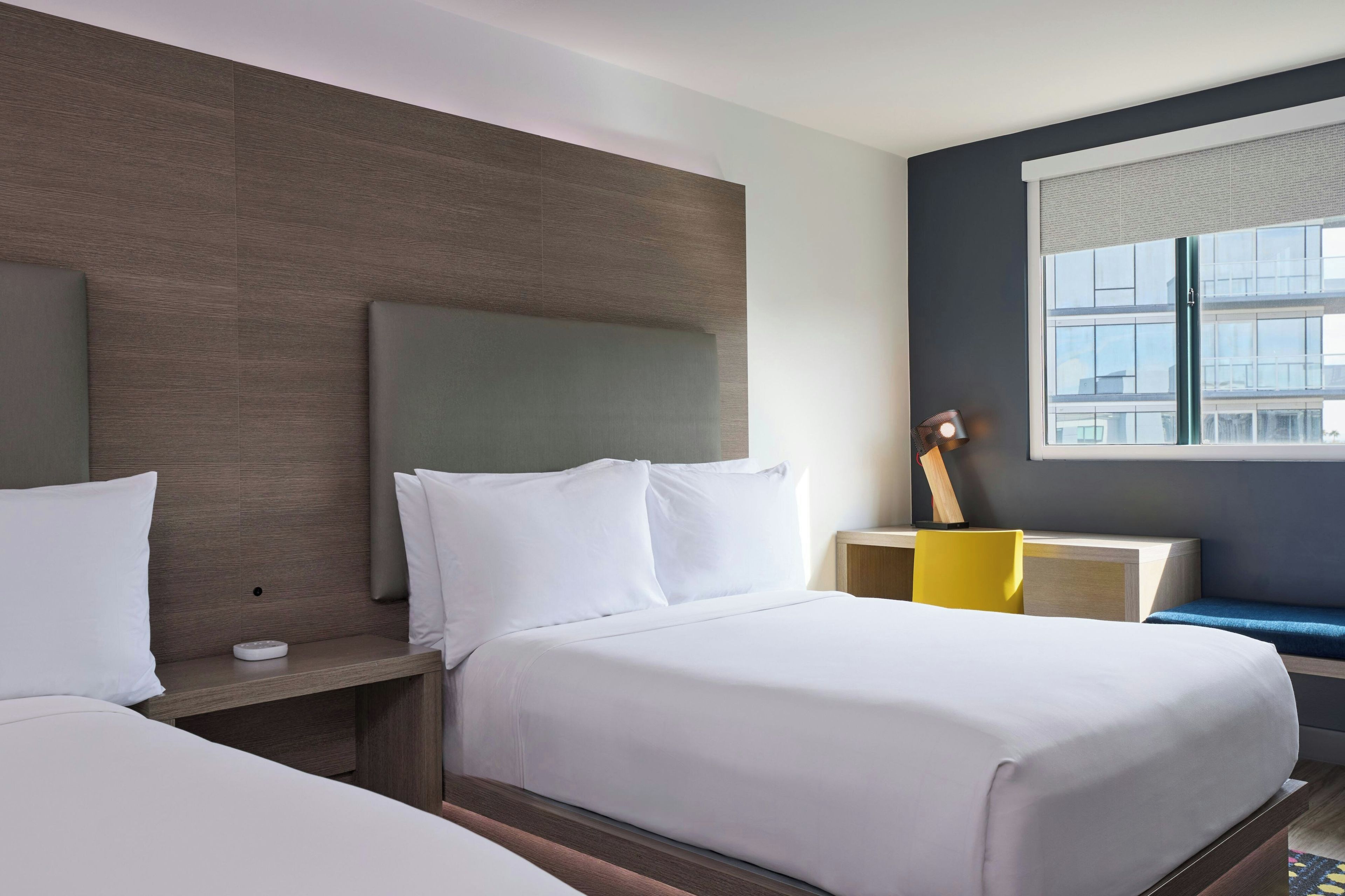 Quarto de hotel com cama, mesas de cabeceira, abajur e janela com vista para prédios.