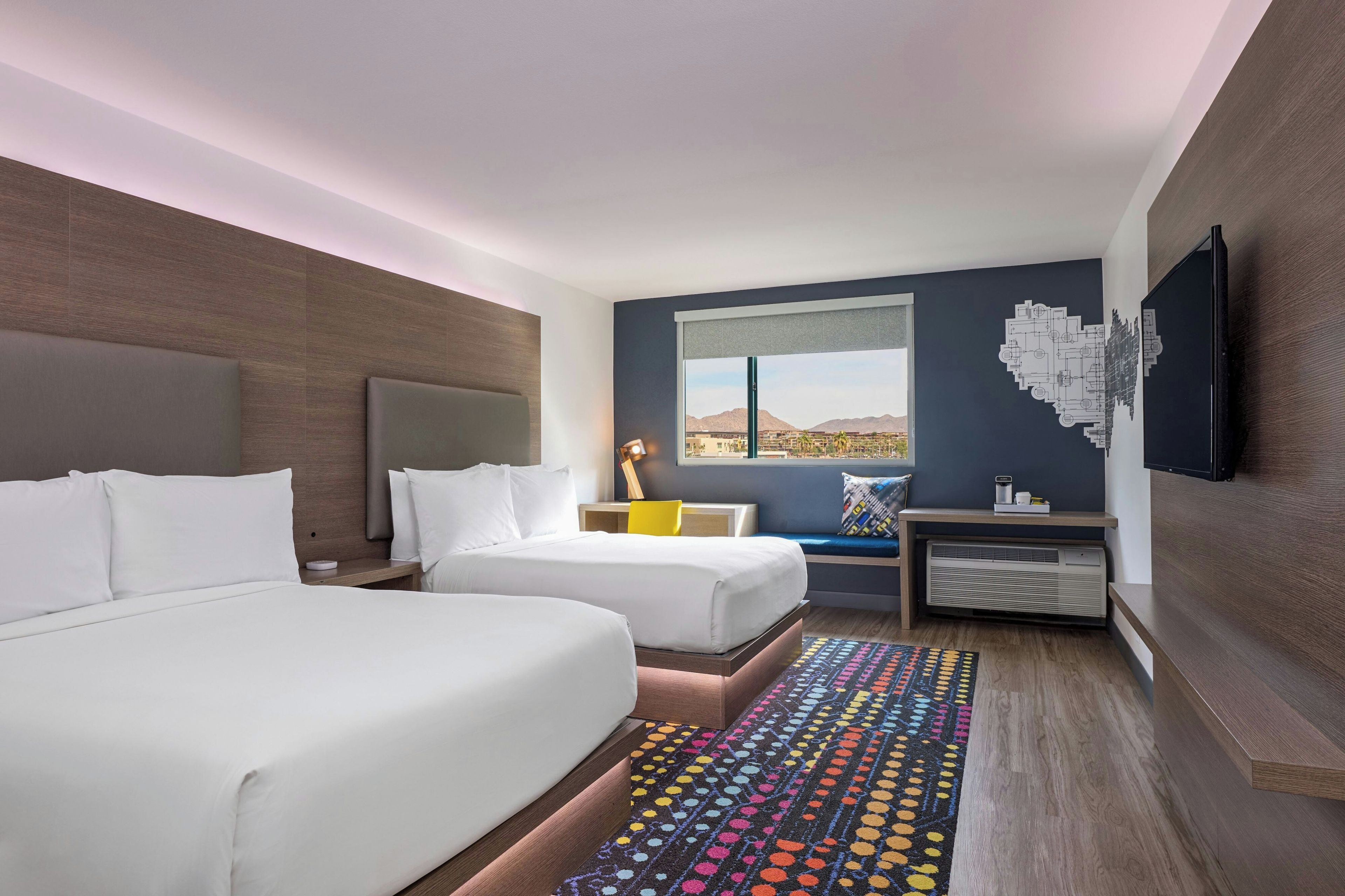 Ein Hotelzimmer mit zwei Betten, einem Schreibtisch, einem Stuhl, einem Fernseher und einem großen Fenster mit Aussicht.