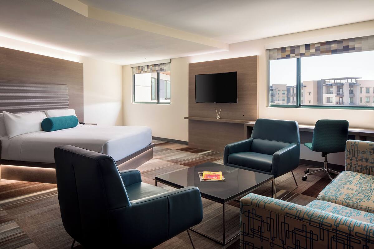 A modern hotel room with a bed, a flat-screen TV mounted on the wall, a seating area with a coffee table, and a large window offering a city view.