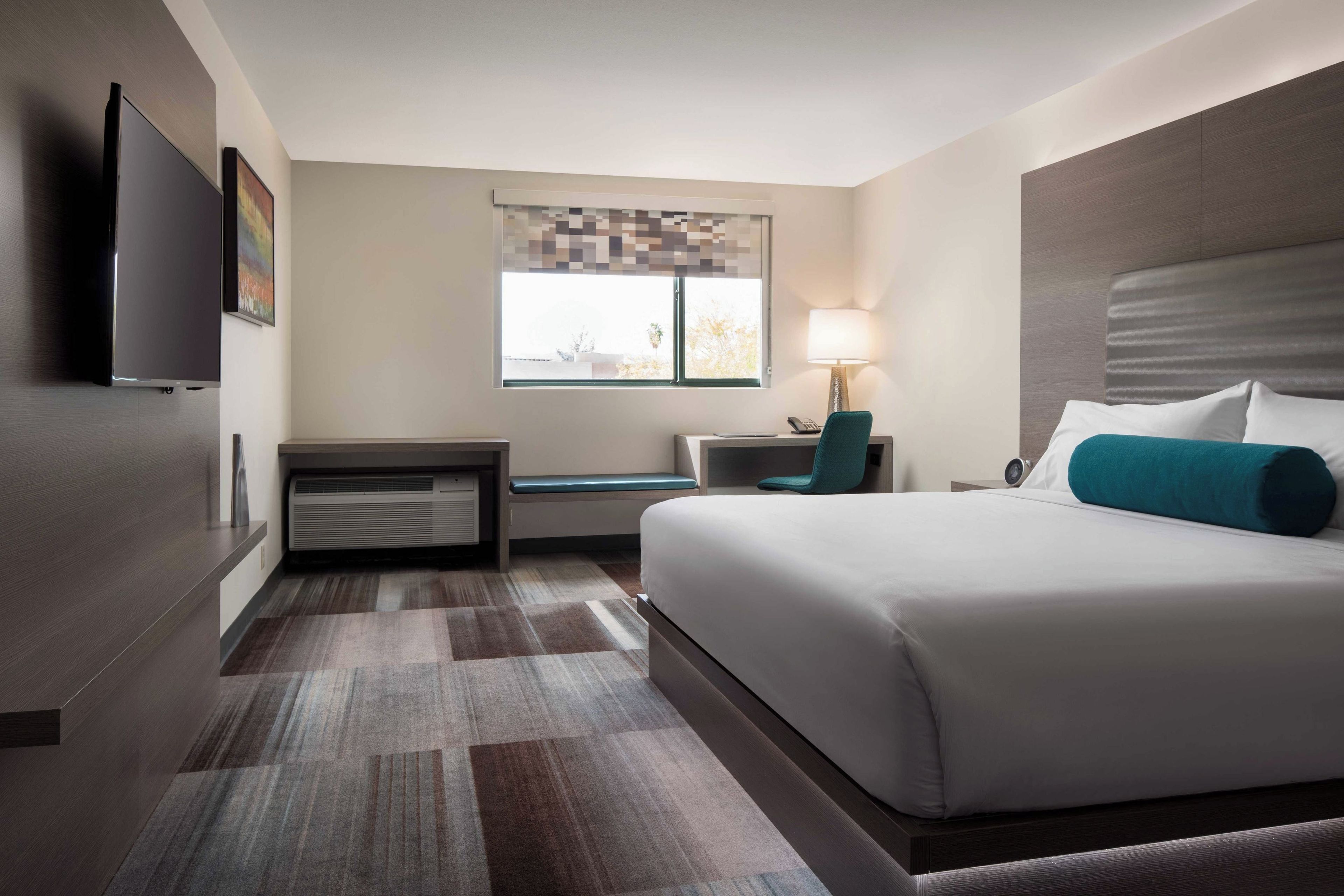 A hotel room with a bed, a desk, a chair, a TV, a window with a view, and a lamp.