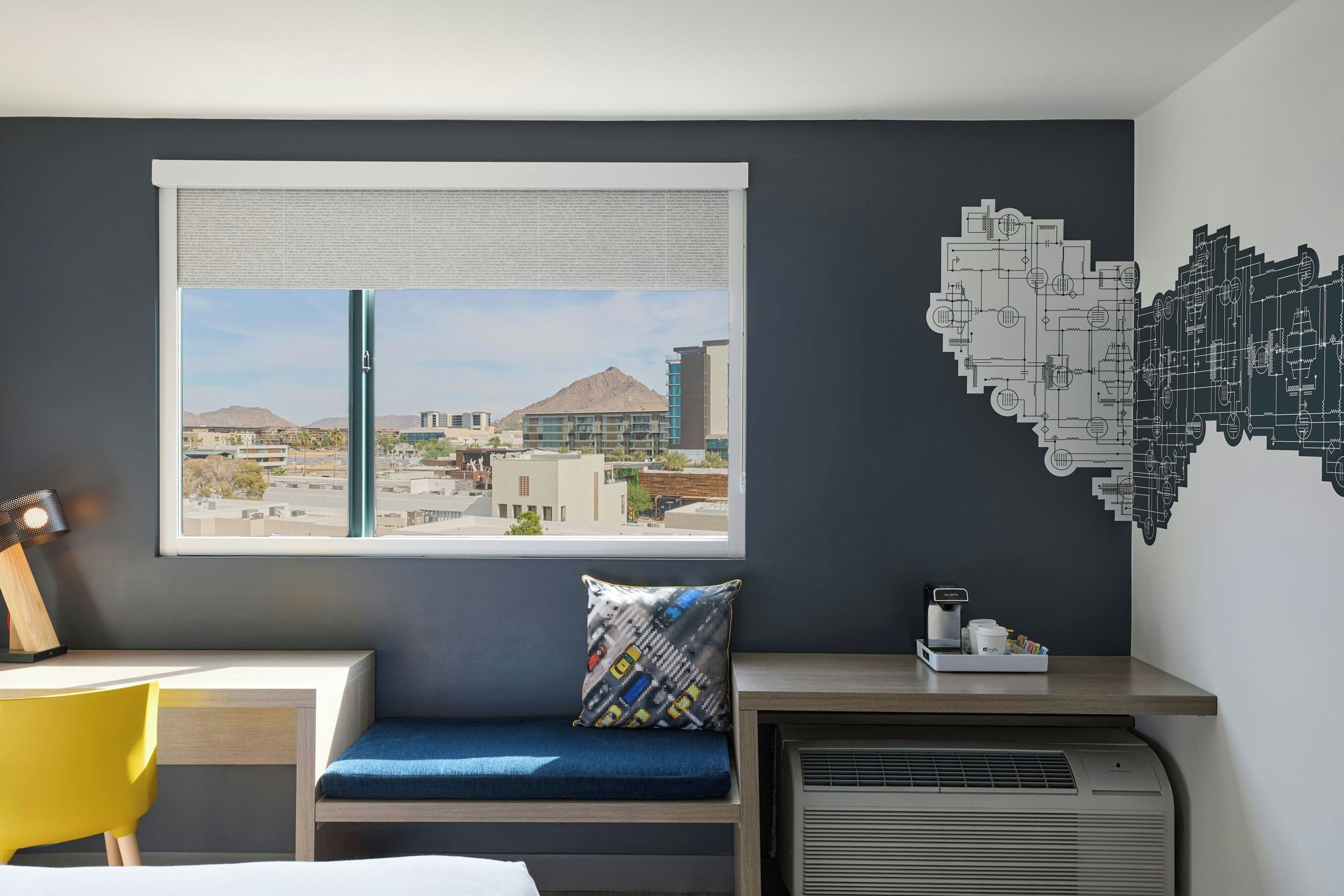 A modern room with a view of a cityscape, a window with blinds, a desk with a lamp, a blue cushioned seat, a wall art of circuit patterns, and a coffee machine.