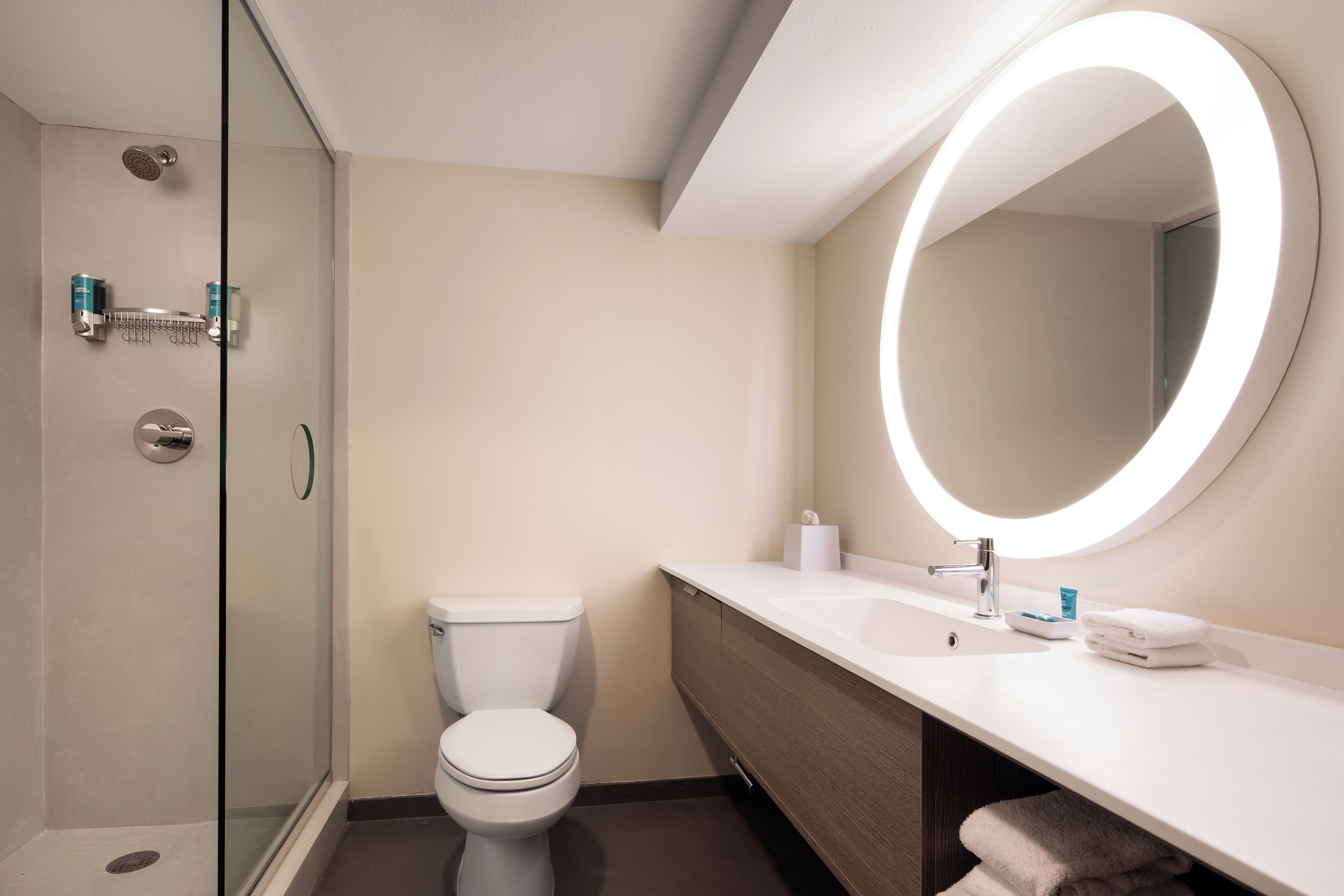 Un baño moderno con cabina de ducha de vidrio, inodoro, tocador con lavamanos y un espejo grande con luz circular.