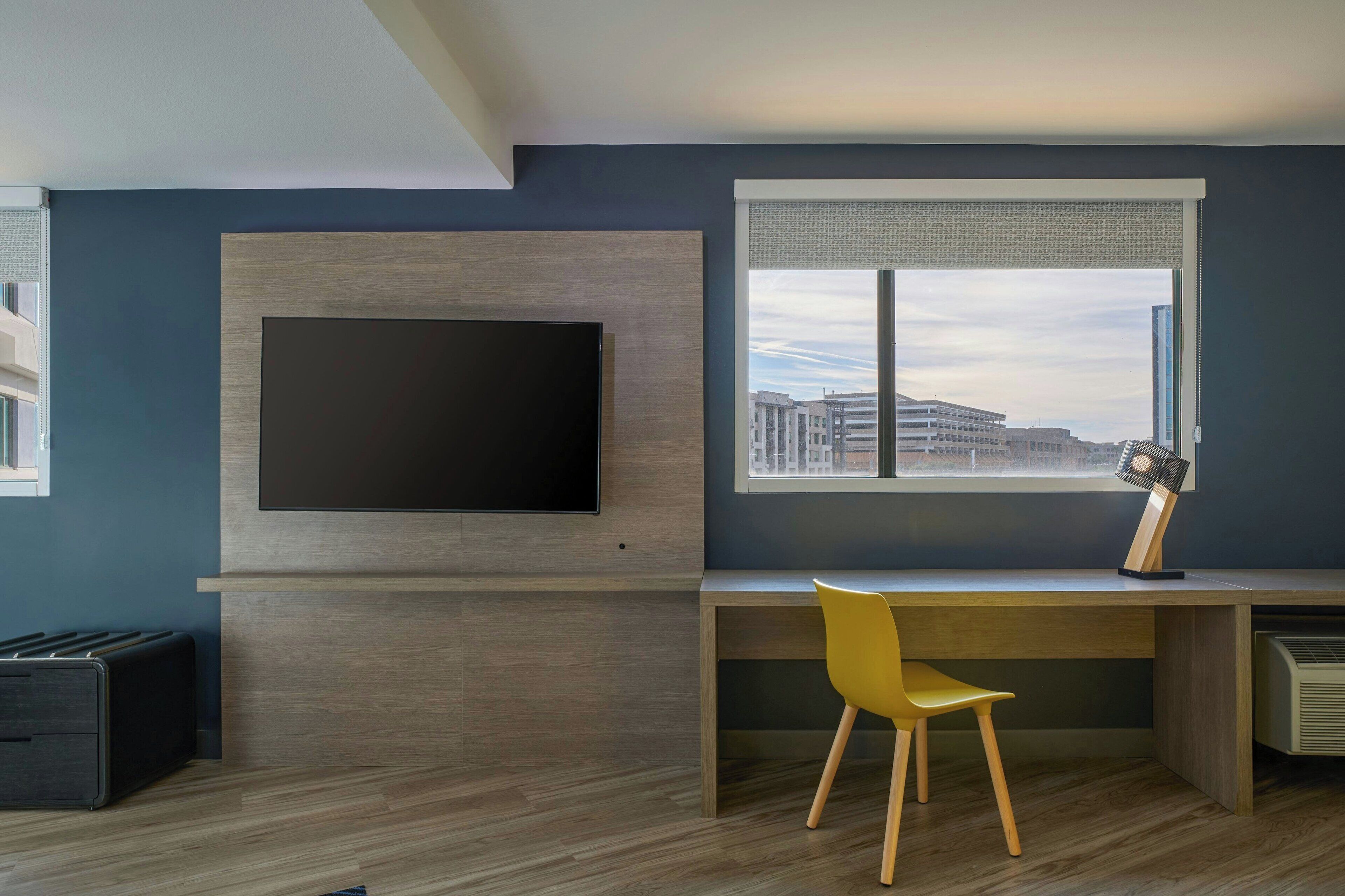 A modern room with a large flat-screen TV mounted on the wall, a desk with a chair, and a window with a view of buildings.