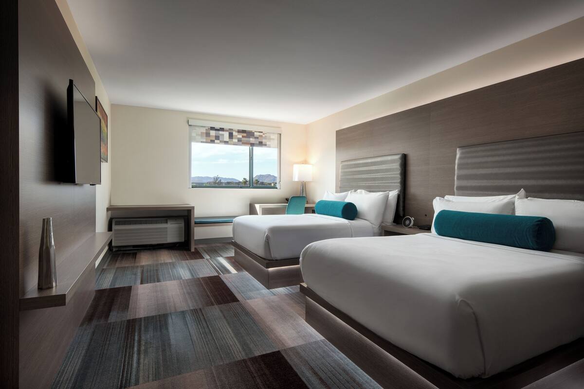 A hotel room with two beds, a desk, a lamp, and a window with a view.