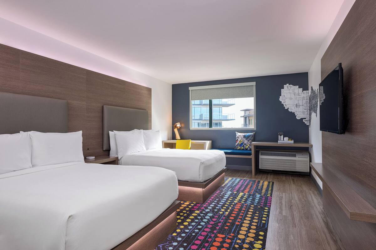 A hotel room with two beds, a large window, a flat-screen TV, and a colorful rug.