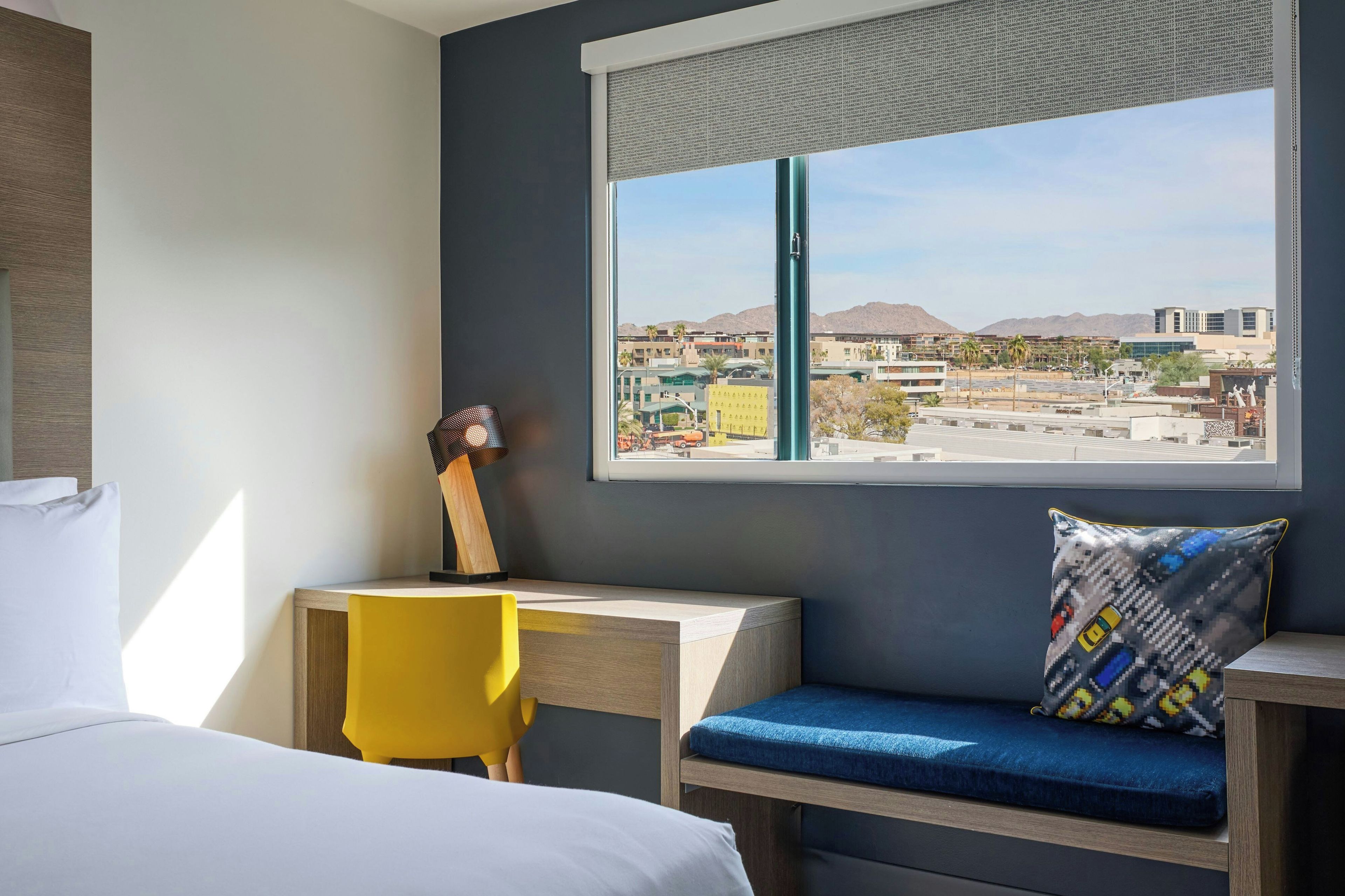 Une chambre d’hôtel avec un lit, un bureau, une chaise, une fenêtre donnant sur un paysage urbain et une guitare appuyée contre le mur.