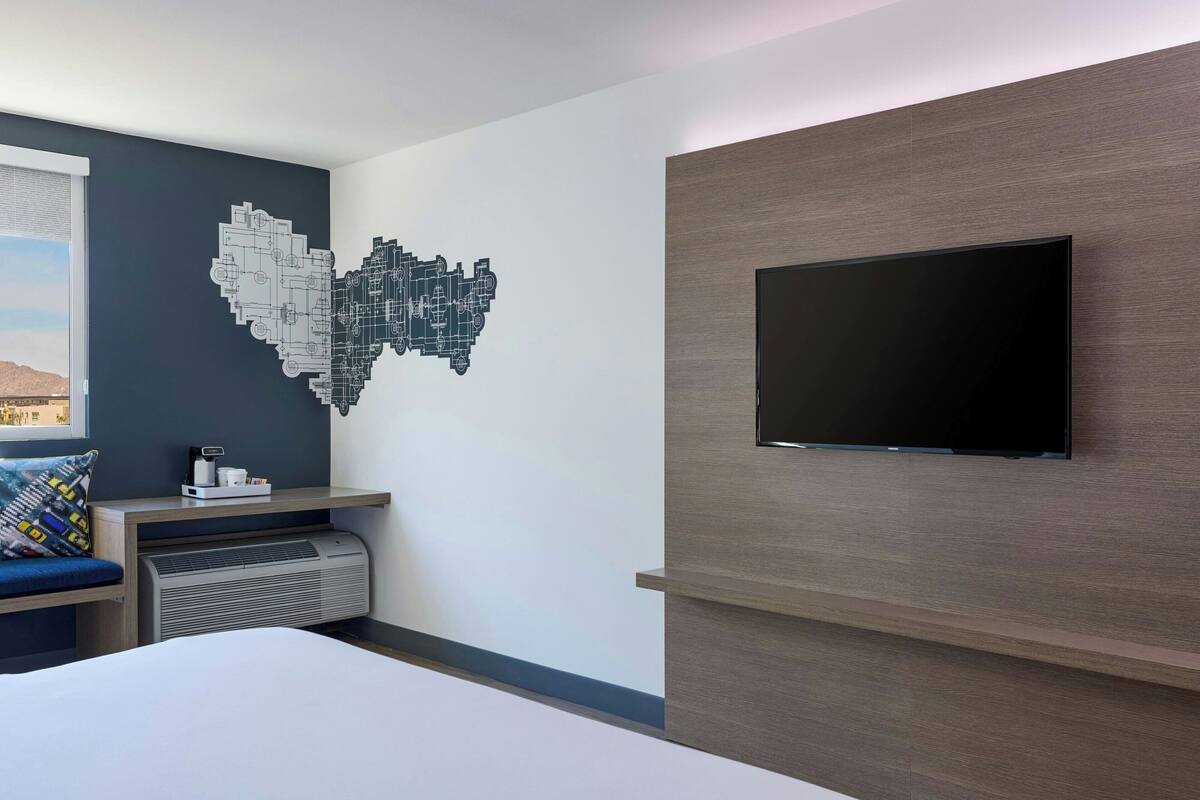A modern hotel room with a large bed, a flat-screen TV mounted on a wooden wall, a wall art piece, a window with a view, and a small table with a coffee maker.