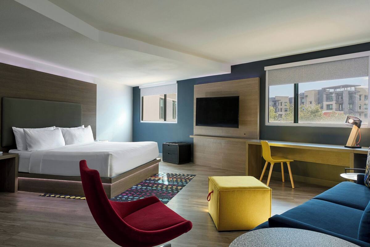 A modern hotel room with a bed, a desk, a red chair, a yellow ottoman, and a blue sofa.