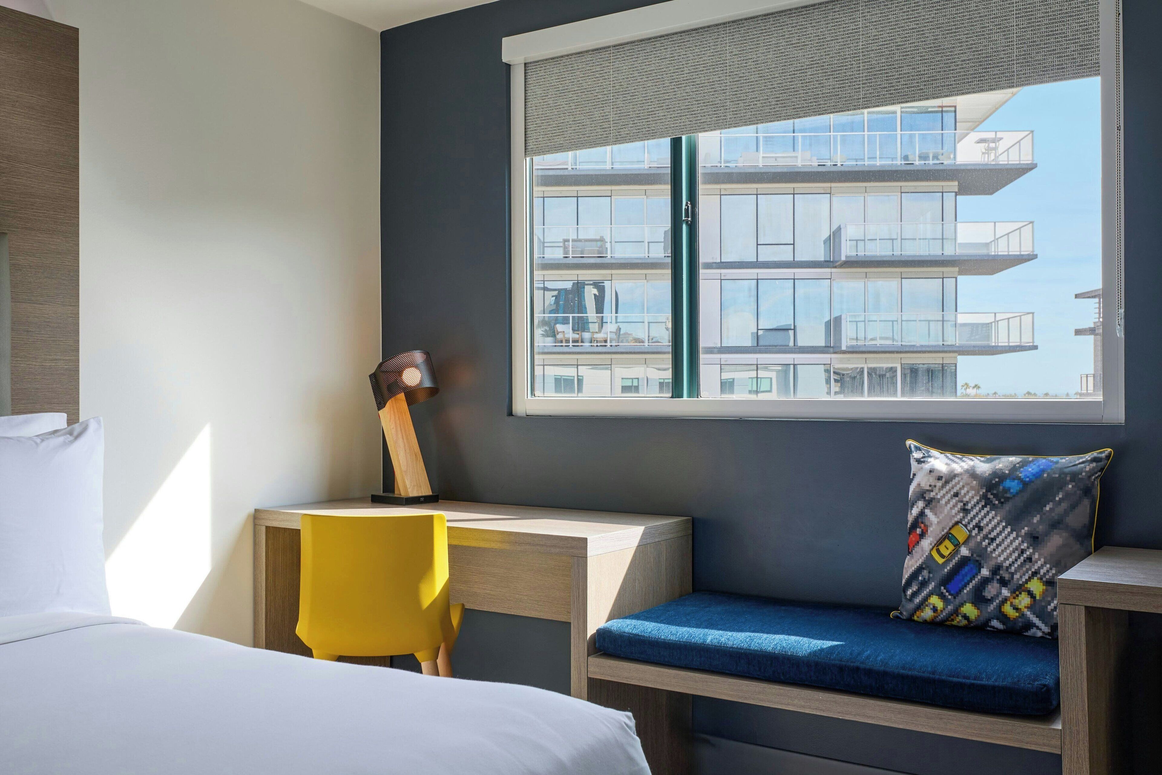 A modern hotel room with a bed, a desk, a chair, and a window with a view of a high-rise building.