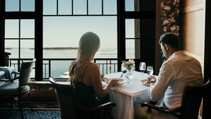 A dining area with a table set for two, large windows offering a view of the water, and a floral wallpaper on the wall. - Oak Bay Beach Hotel (Victoria)